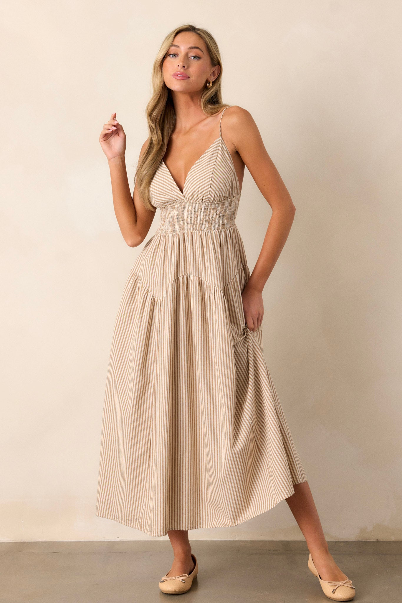 Movement view of this tan midi dress that features a deep v-neckline, a smocked waistband, functional hip pockets, a stripe print, and adjustable spaghetti straps.