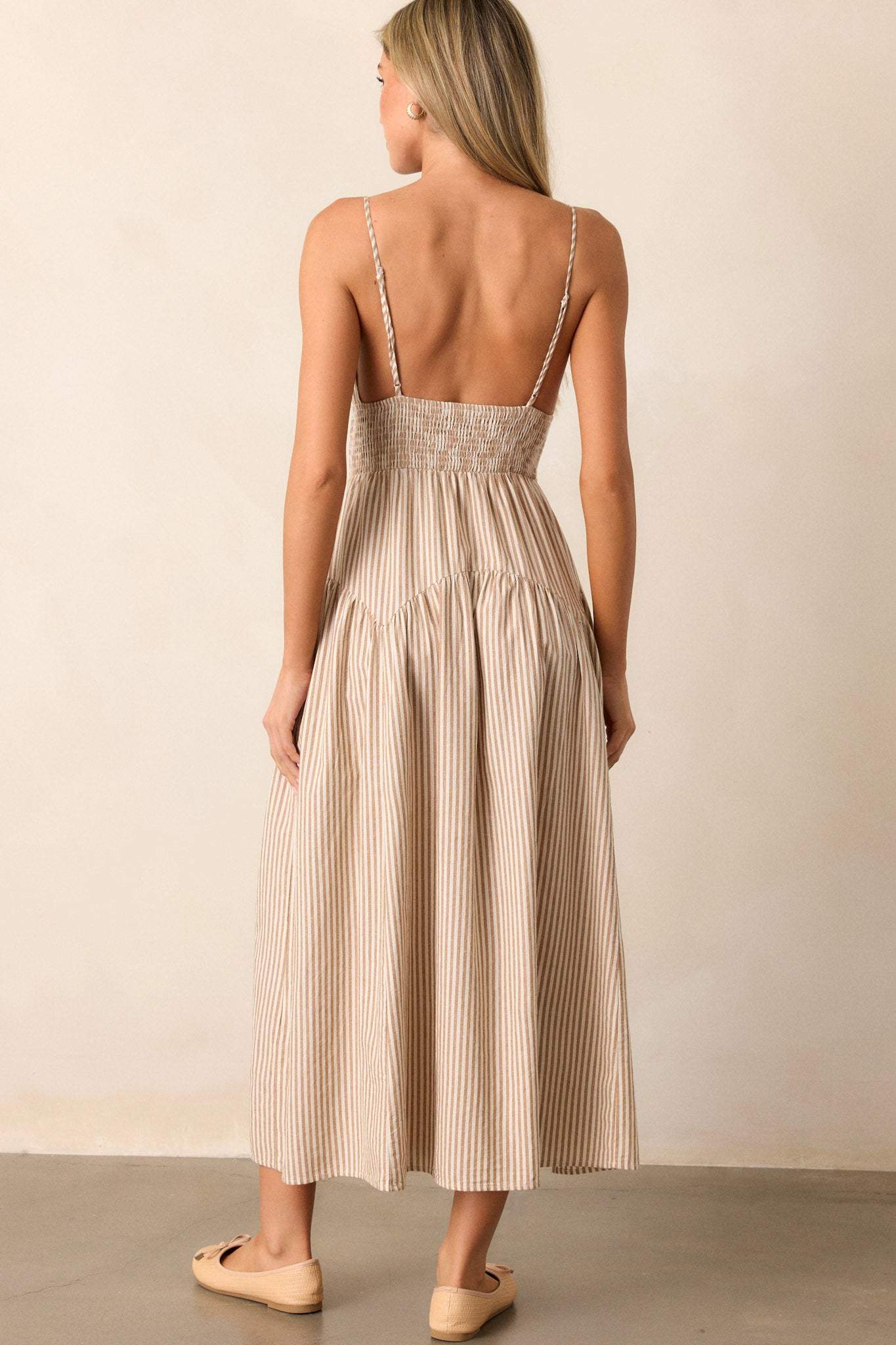 Back view of this tan midi dress  that features a deep v-neckline, a smocked waistband, functional hip pockets, a stripe print, and adjustable spaghetti straps.