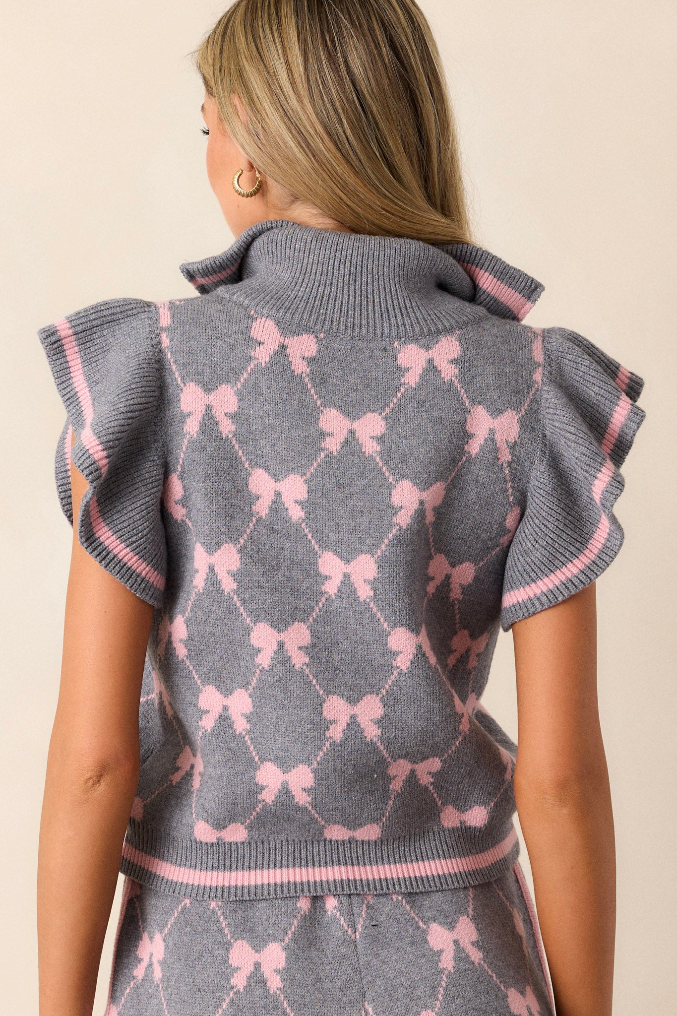 Back view of a grey top featuring a functional quarter zip-up front, a collared neckline, a pink bow print, flutter sleeves, and a soft knit fabric.