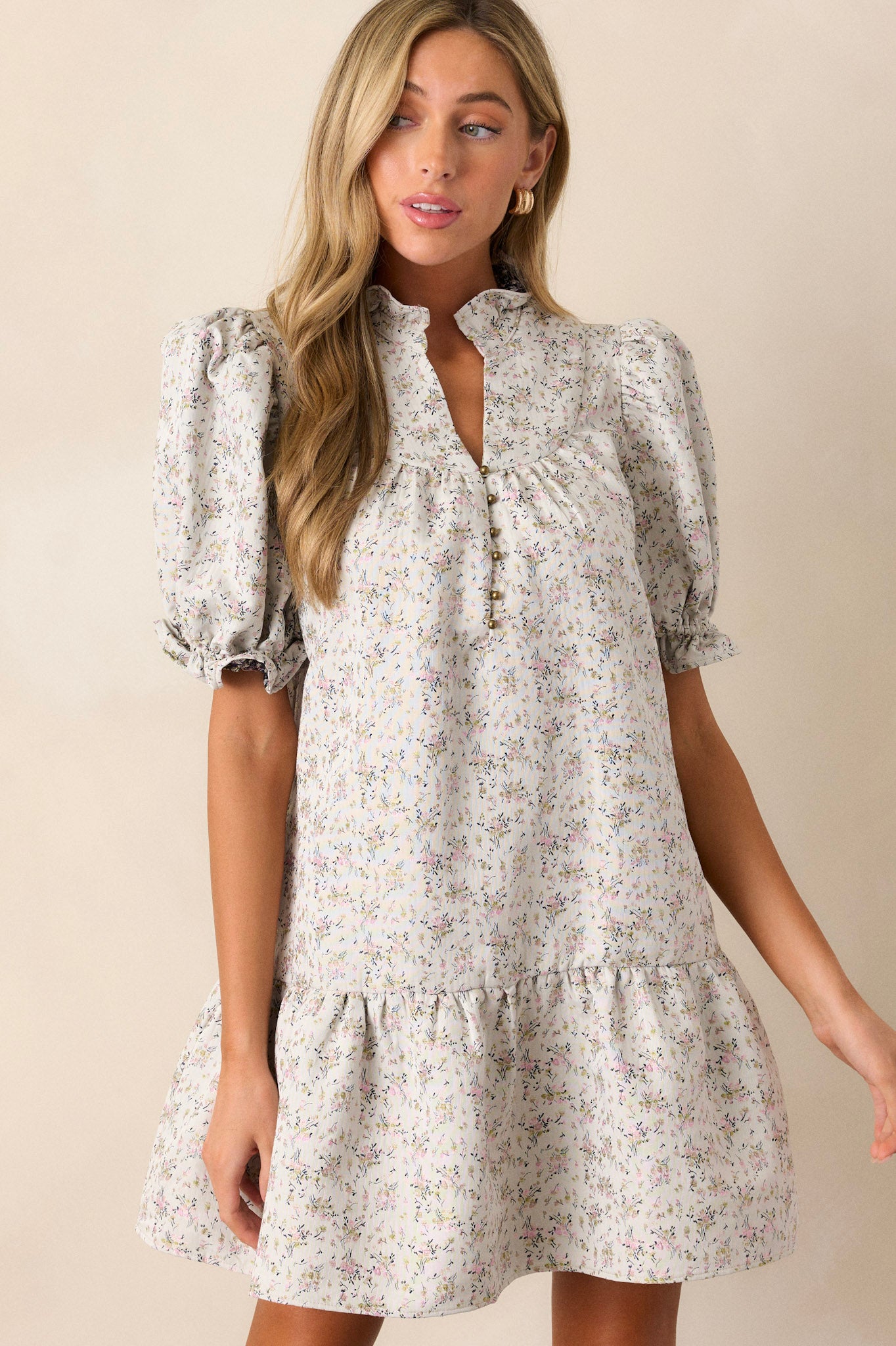 Front view of this almond floral dress that features a ruffle collared neckline, a split neckline with a functional button up bust, a soft quilt texture, a small floral pattern, a tiered design, a ruffled trim and short elastic cuffed puff sleeves.