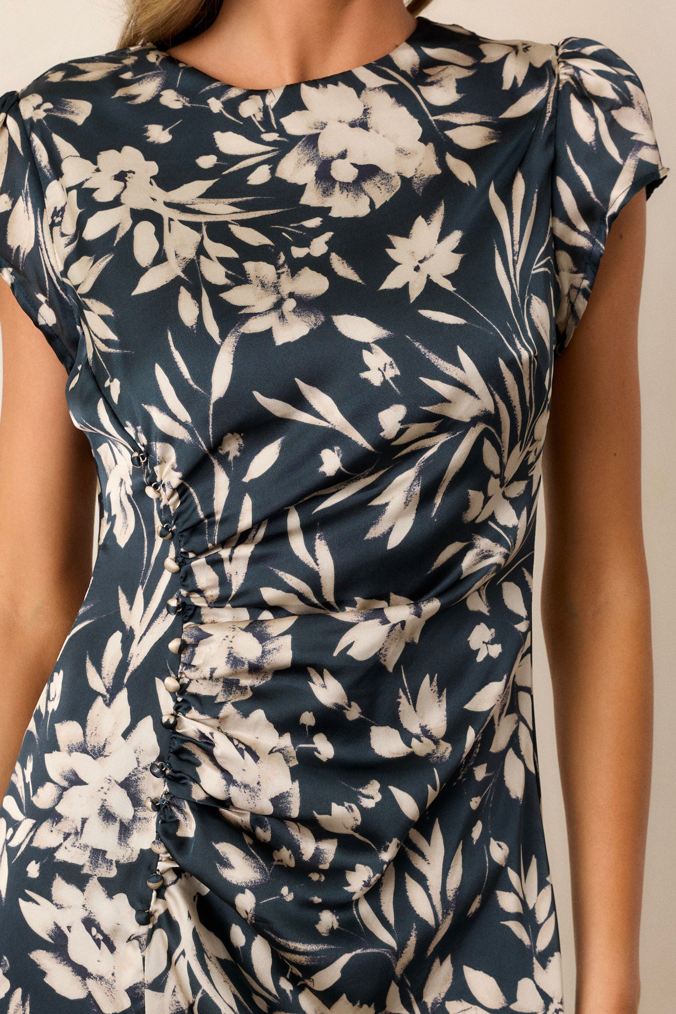 Close-up view of this dark teal floral dress that features a high neckline, a non-functional button design on the bust to the waist, an off-white floral design, a leg slit, a discrete back zipper, a button and loop closure, and short sleeves.