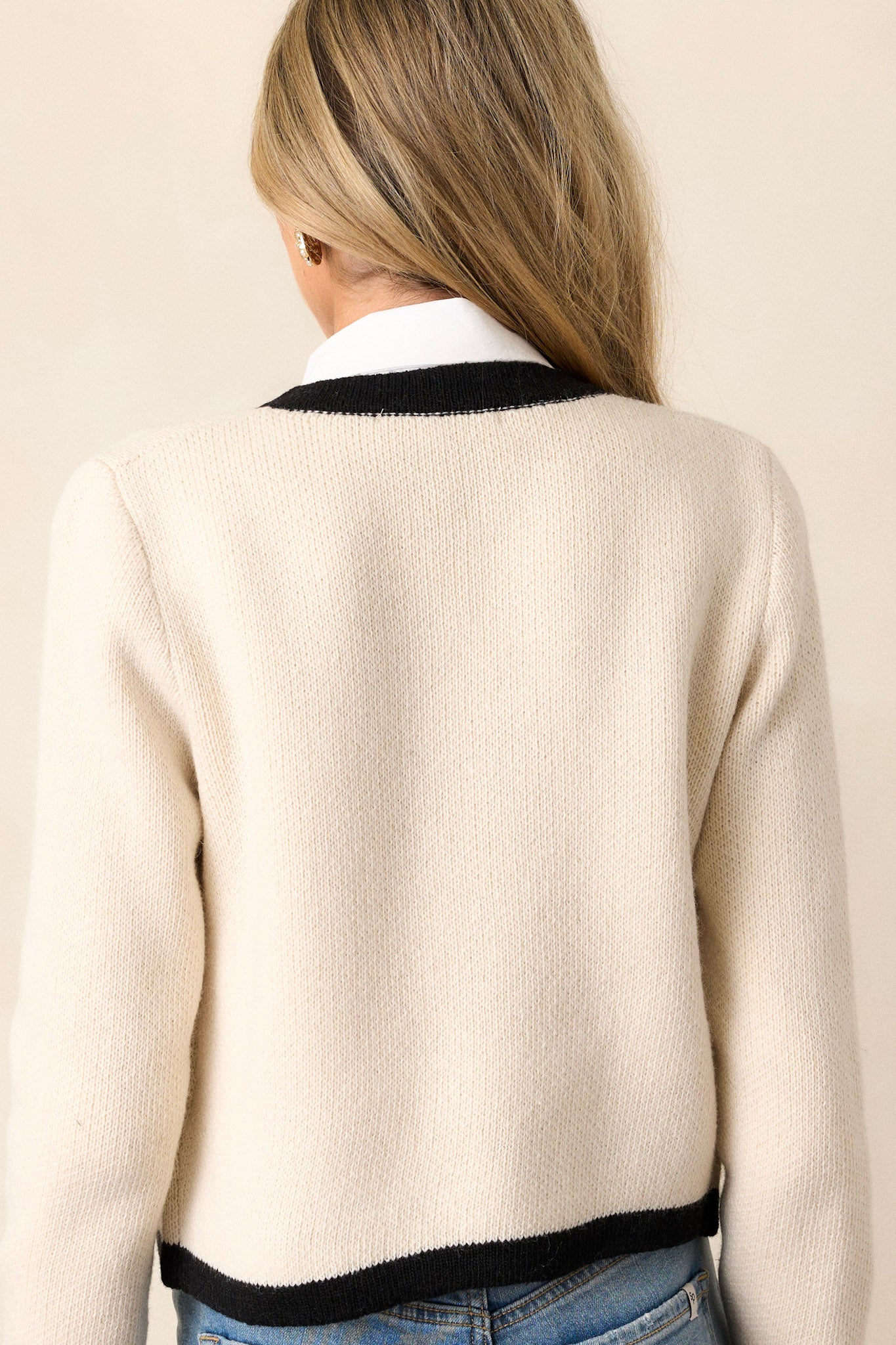 Back view of an ivory cardigan that features a black contrast trim, functional gold buttons down the front, two functional pockets, and a cozy knit fabric.