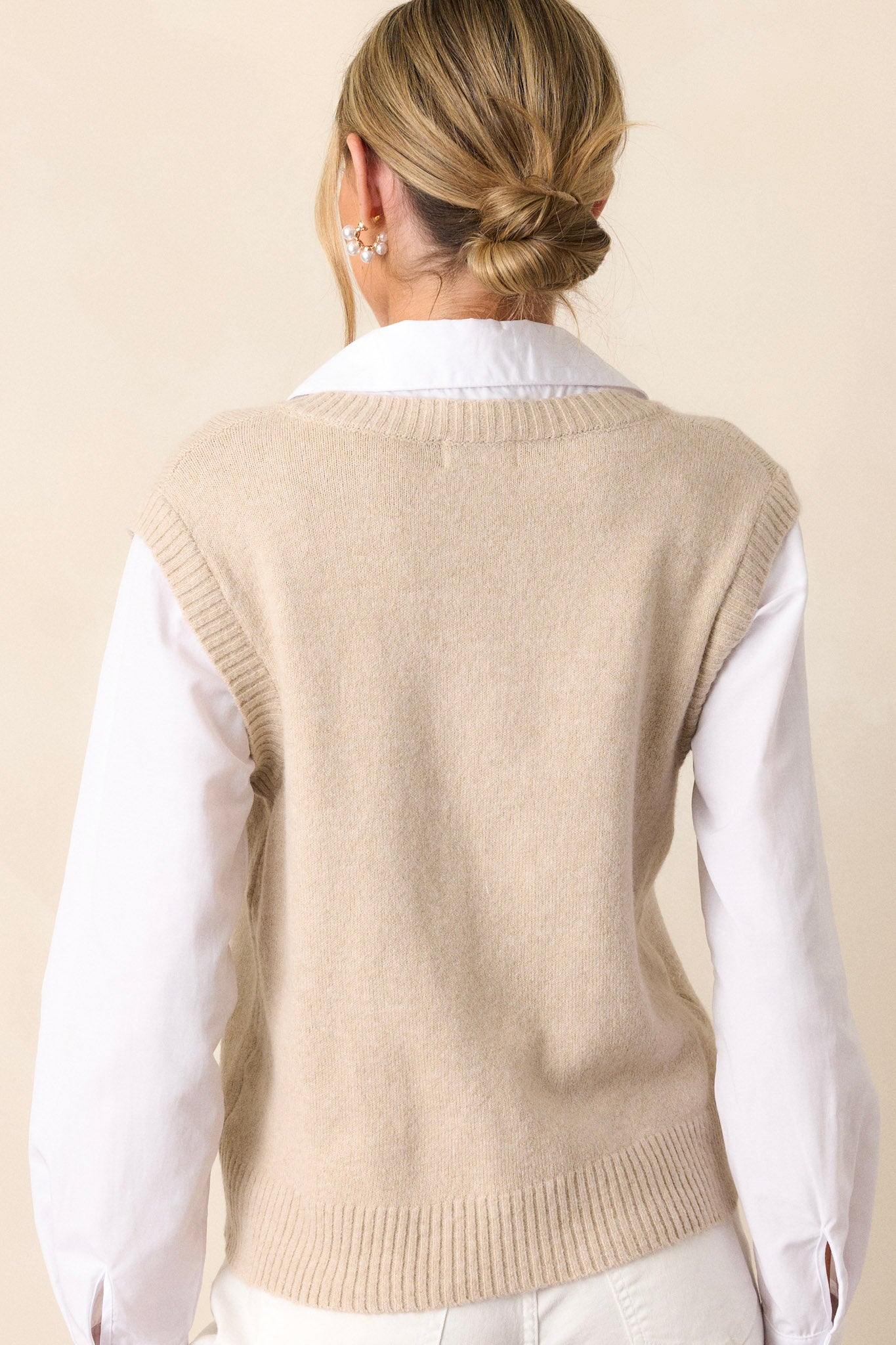 Back view of this beige sweater that features a white collared neckline, a functional button neckline, a layered beige sweater vest, a relaxed fit and long white sleeves.