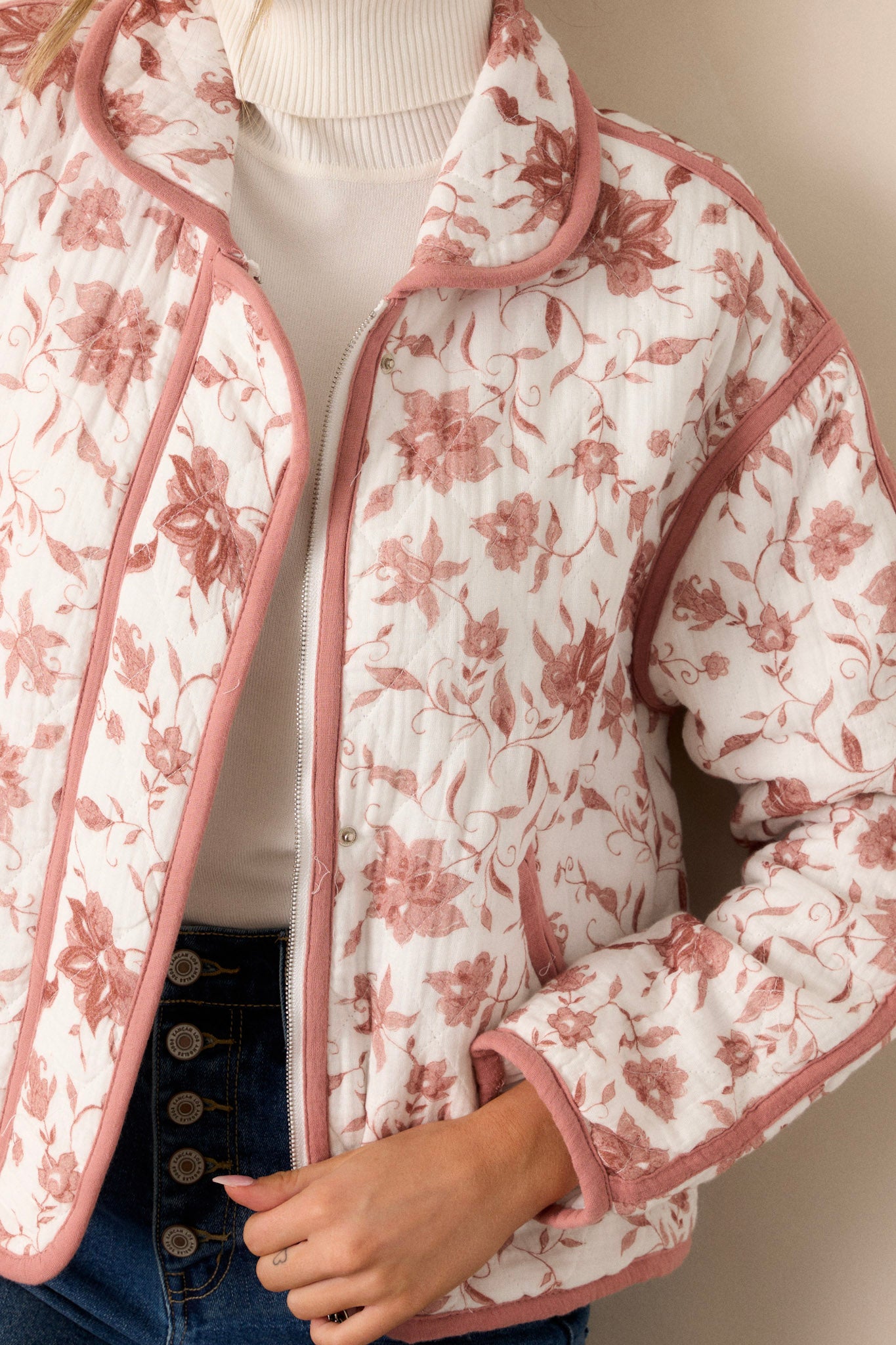 Close-up of this dusty pink jacket that features a collared neckline, a full zip front, a solid dusty pink trim, an ivory floral print, a soft quilted material and snap buttons for extra support.