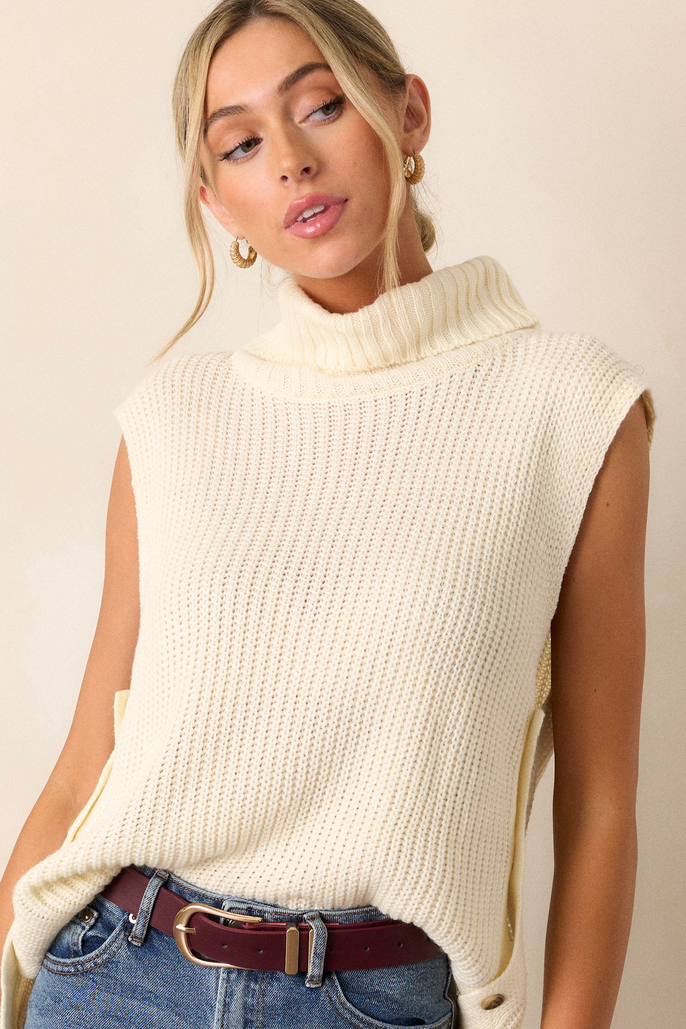 This ivory sweater features a turtleneck, a ribbed texture, multiple buttons on side, and a sleeveless design.