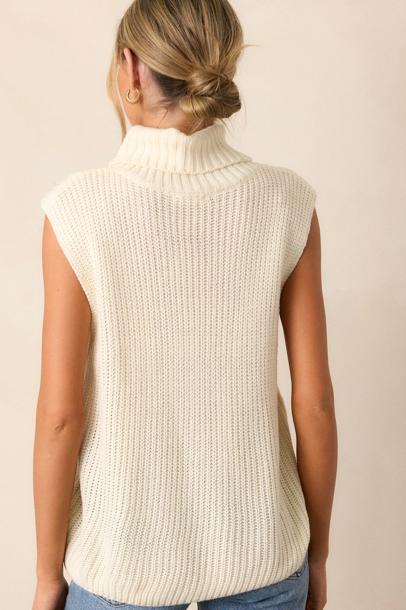 Back view of this ivory sweater that features a turtleneck, a ribbed texture, multiple buttons on side, and a sleeveless design.