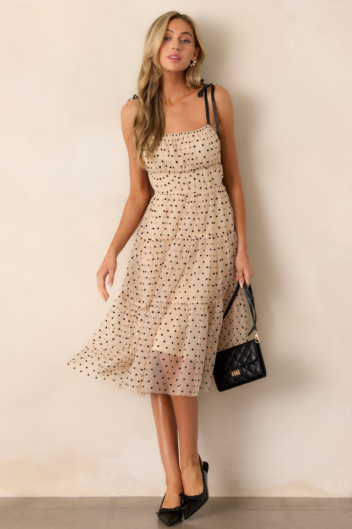 Front angled view of a beige midi dress featuring a square neckline, thin self-tie straps, an elastic waistline, tiers, black polka dots, and tulle