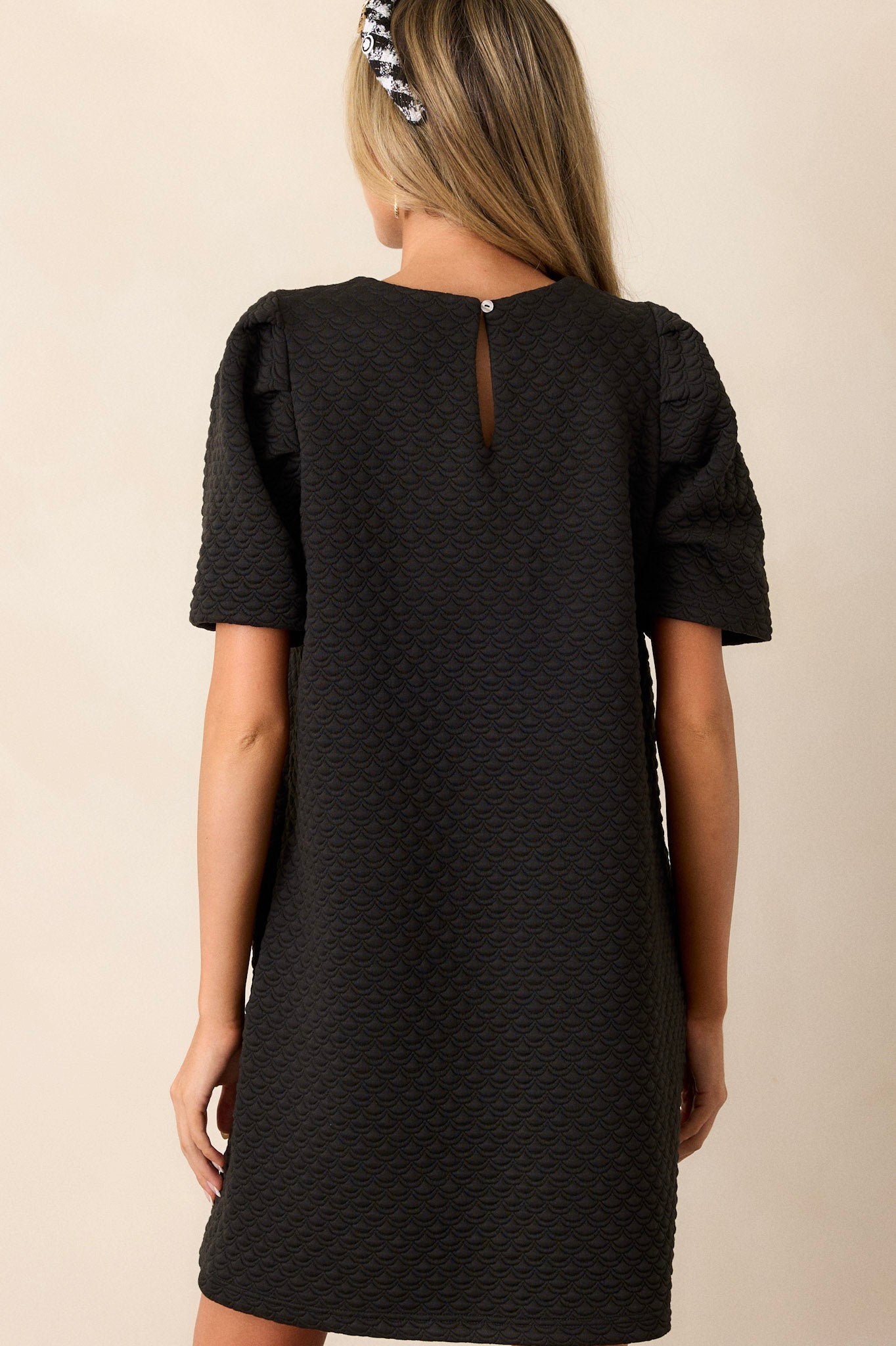 Back view of this black dress featuring a crew neckline, rhinestone embellishments around neckline, puff sleeves, a textured material, and a back cutout with loop closure.