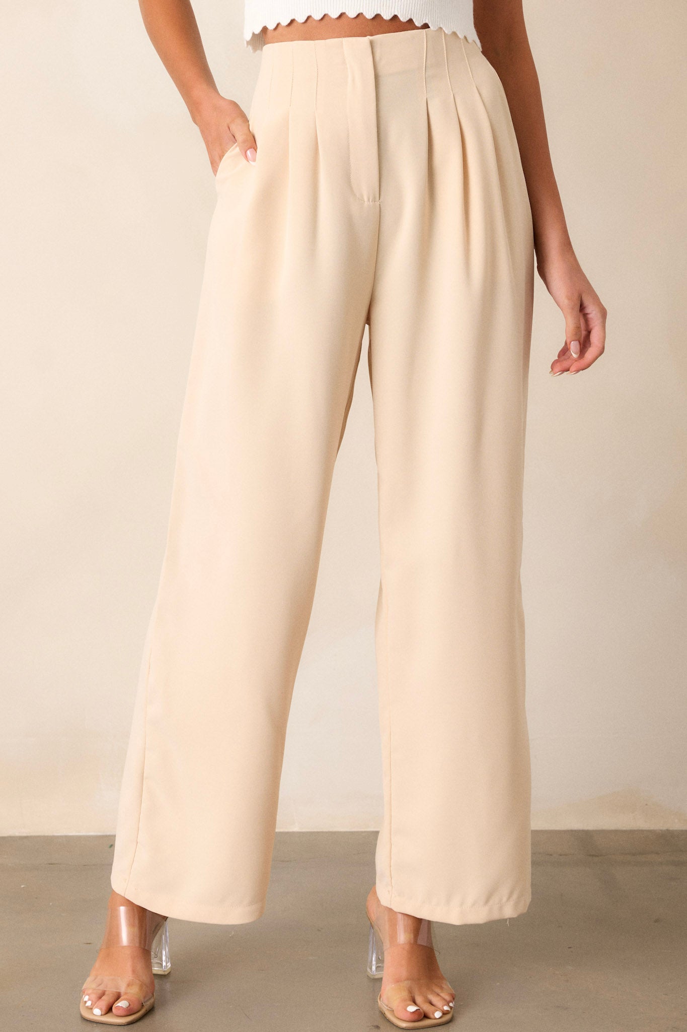 Full-length view of beige pants featuring a high-waisted design, fitted waistband, slight pleats, functional hip pockets, and a wide leg silhouette