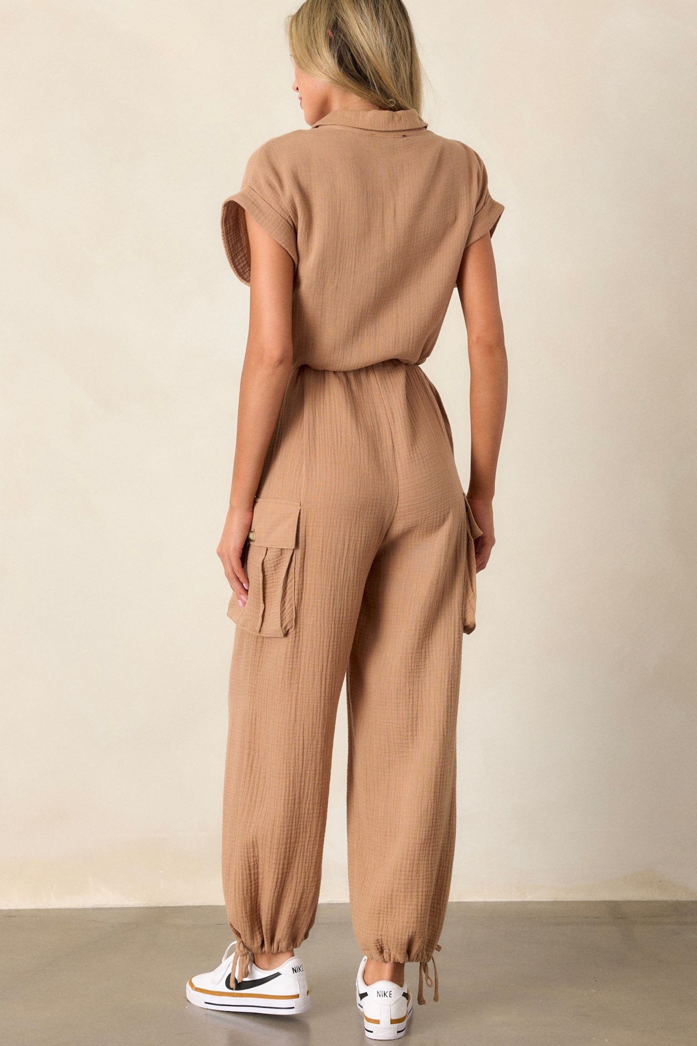 Back view of the camel jumpsuit highlighting the continuation of the collared neckline, the self-tie drawstring waist, and the tapered leg design with drawstring ankles.