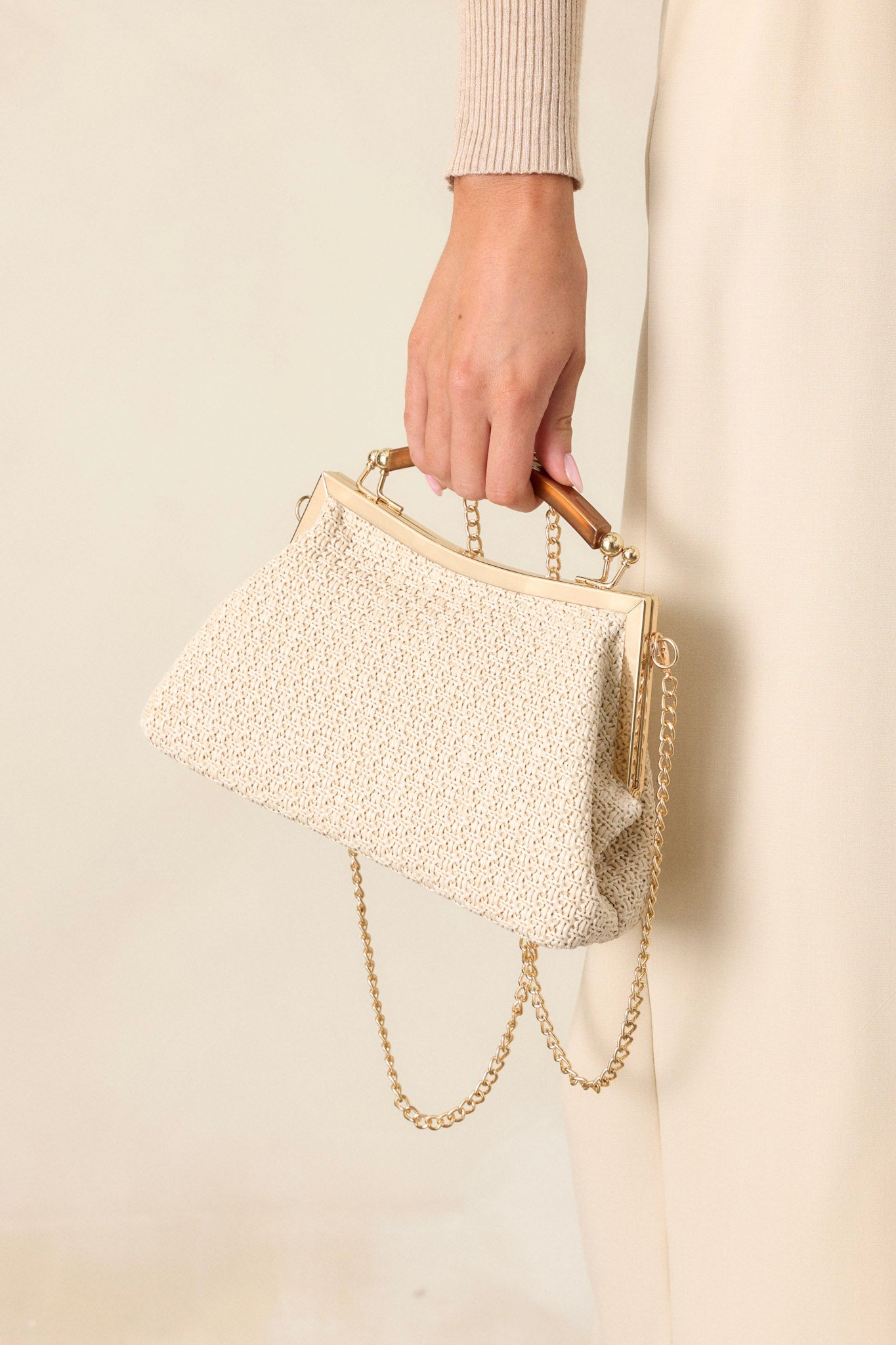 Close-up view of this natural handbag that features a metallic gold handle, gold hardware trim, gold loops for the optional chain, kiss-lock clasps on both sides and a beige woven material.