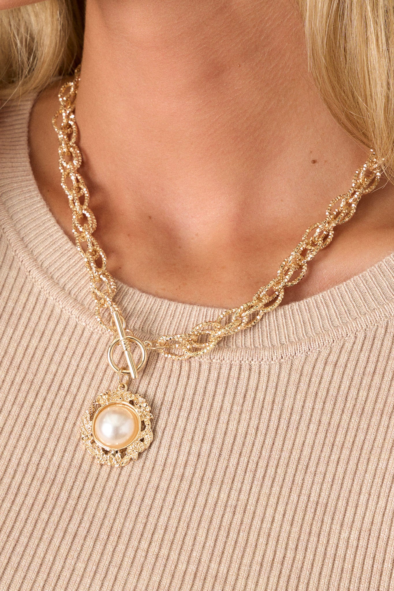 This gold chain necklace features a gold textured chain, a gold pendant with a faux pearl and writing on the back that says "Live in the moment", the pendant is surrounded in gold floral hardware and a toggle clasp closure.