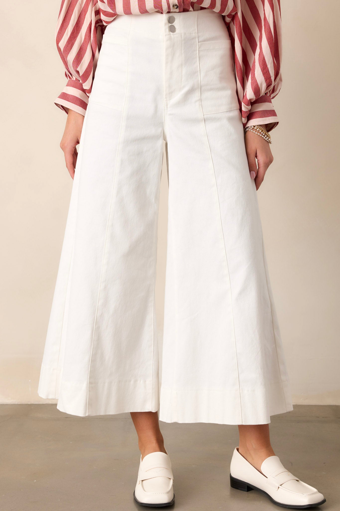 Front view of white pants featuring a high waisted design, a double button closure with a zipper, functional front pockets, front & back seams, and a wide leg with a cropped length.