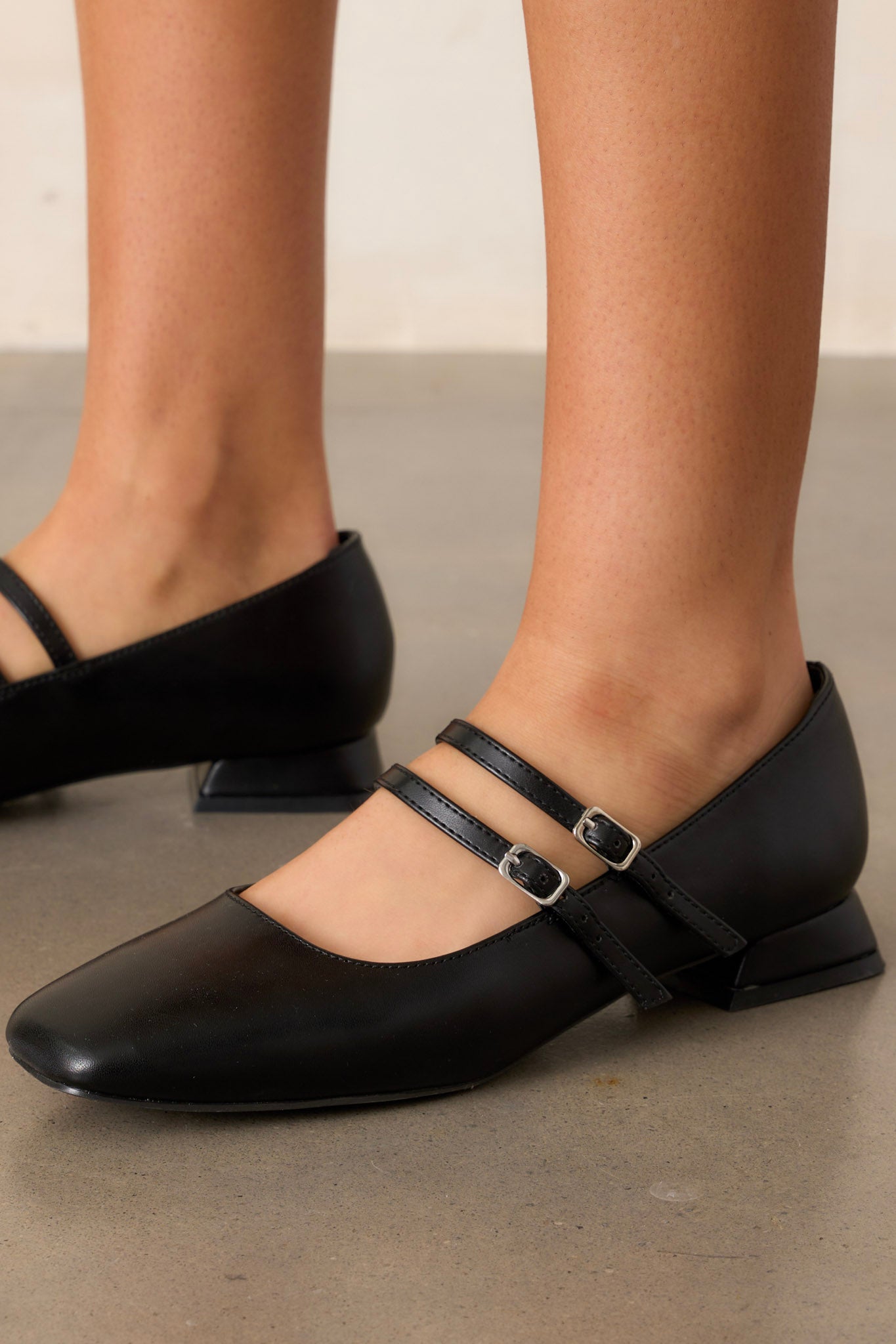 Close-up of these black flats that feature a square toe, two foot straps with silver buckles, a small square heel and a matte black finish.