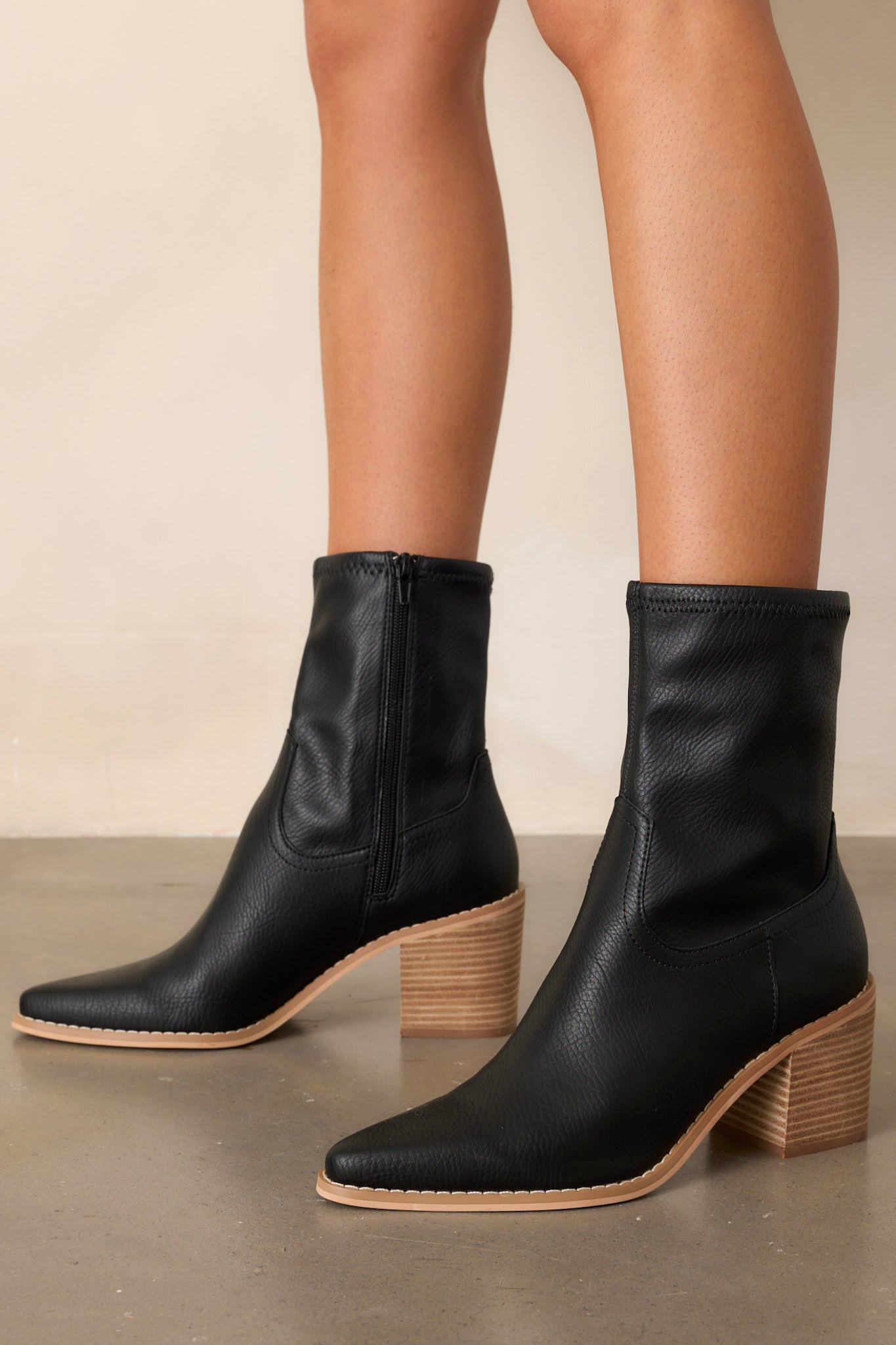 Angled side view of these black boots that feature a pointed toe, a platform heel, a discrete side zipper and a tan sole.