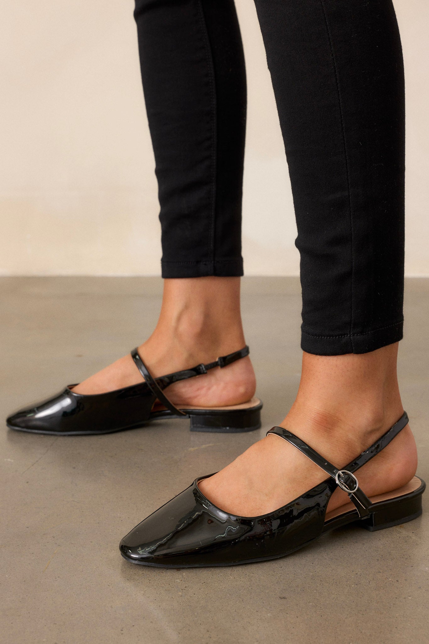 Close-up of these black flats that feature a slightly squared toe, an across the foot strap with a silver buckle, an ankle strap, a small platform, and a shiny black finish.