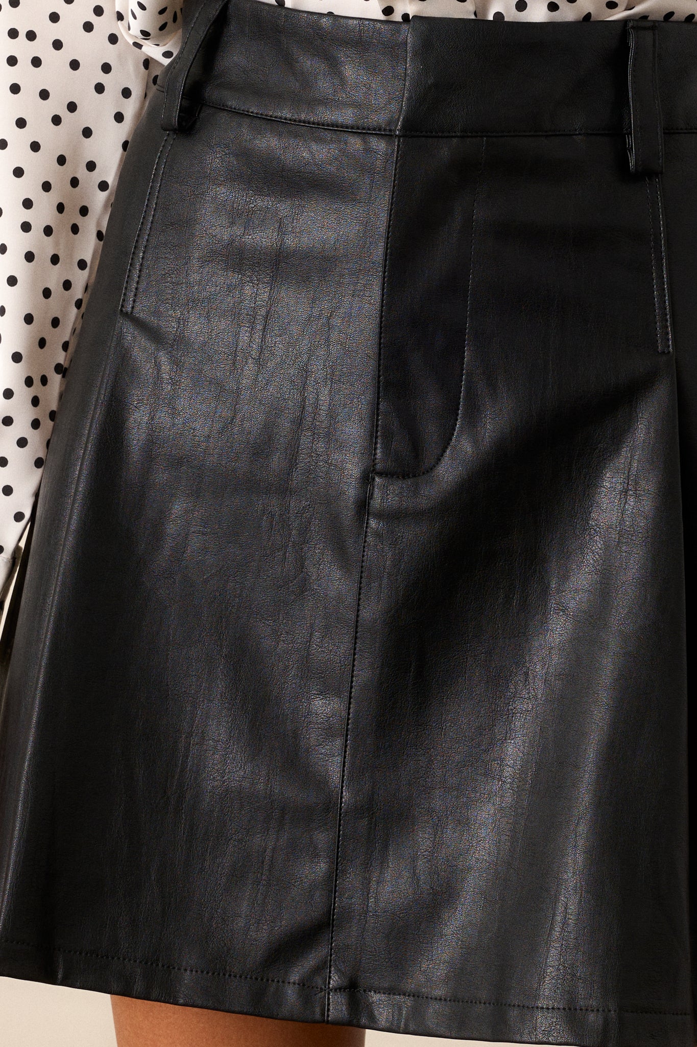 Close-up detail shot highlighting the texture of the faux leather fabric and the stitching along the pleats of the black mini skirt.