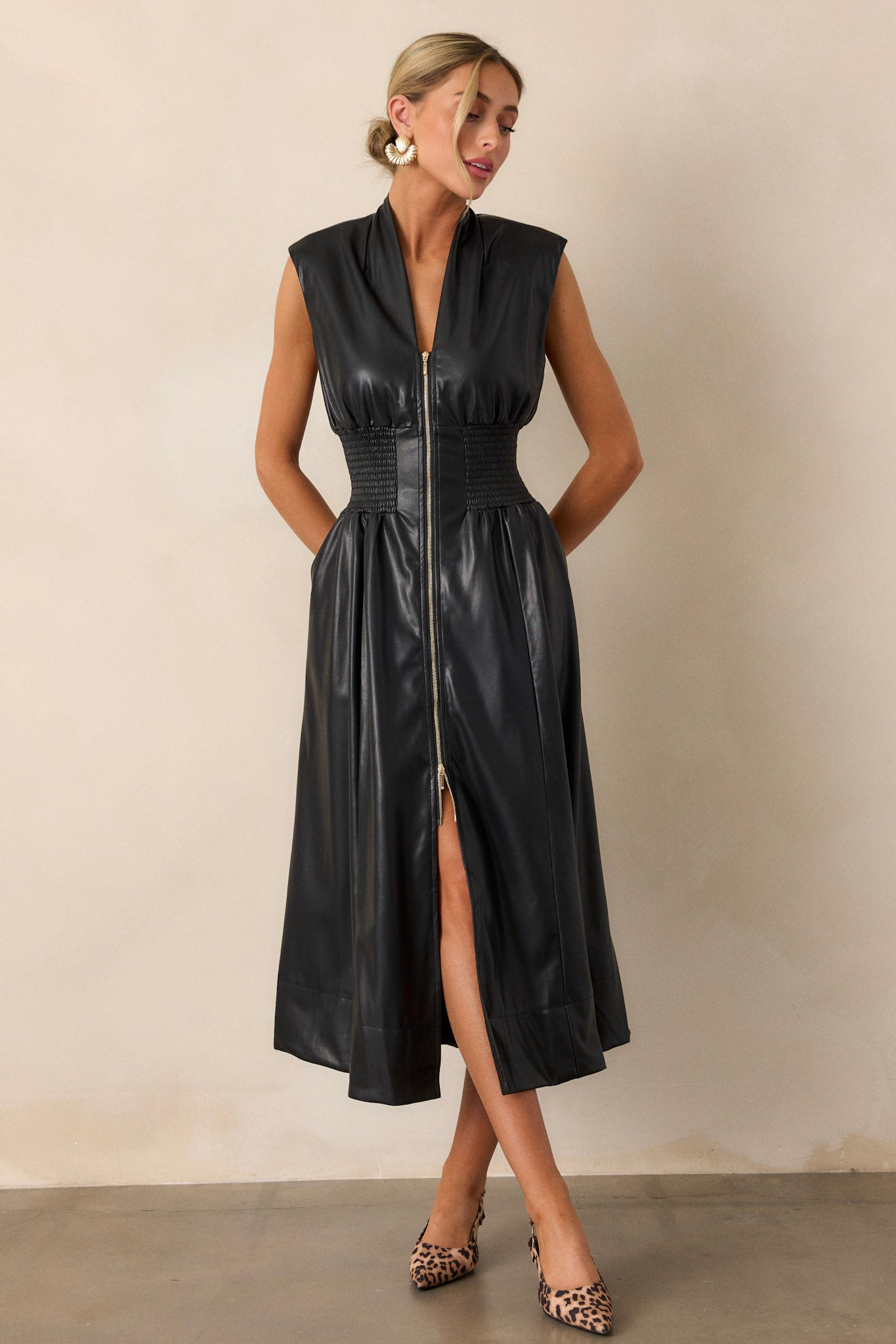 Full view of the black faux leather midi dress featuring a v-neckline, padded shoulders, and a fully smocked waist, highlighting its structured silhouette.