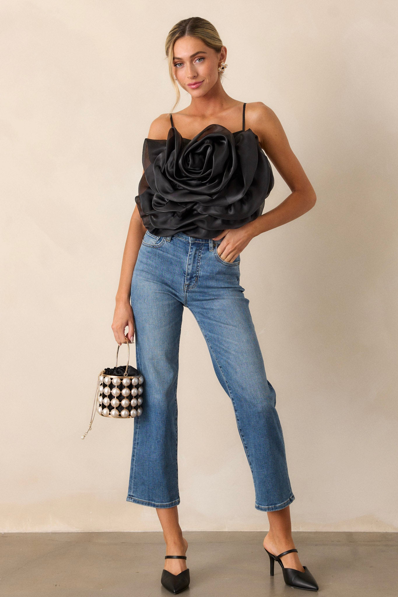 Full length view of a black crop top with a square neckline, thin and stretchy adjustable straps, a fully smocked back, a large 3-D flower, and a cropped hemline