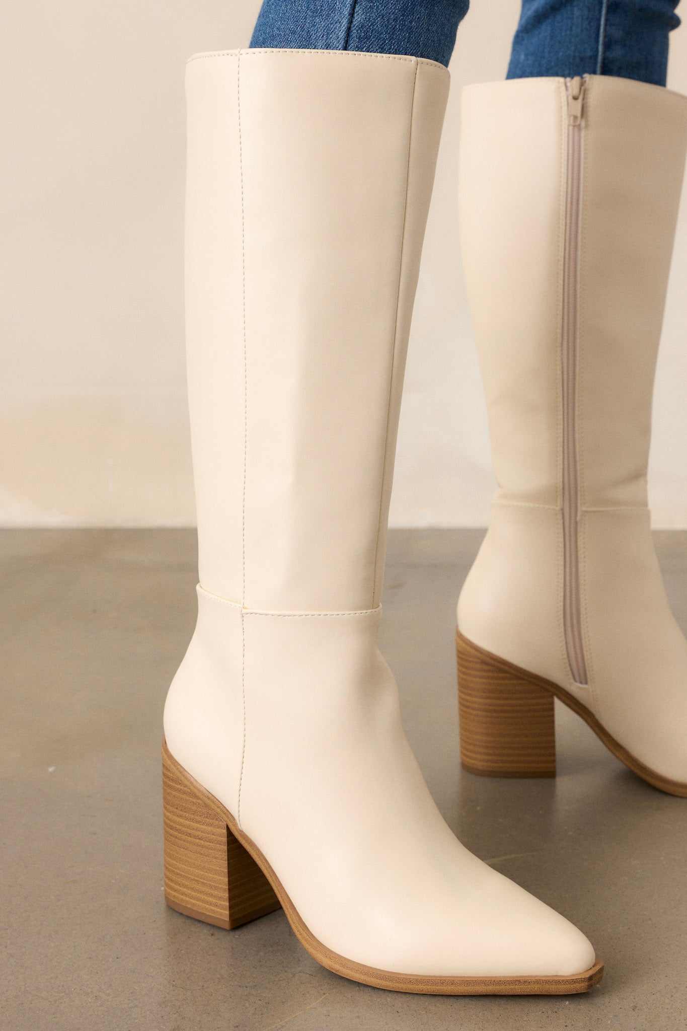 Full view of ivory boots featuring a pointed toe, a sleek faux leather ivory material, a brown block heel, a functional side zipper, a knee high design and a brown sole.