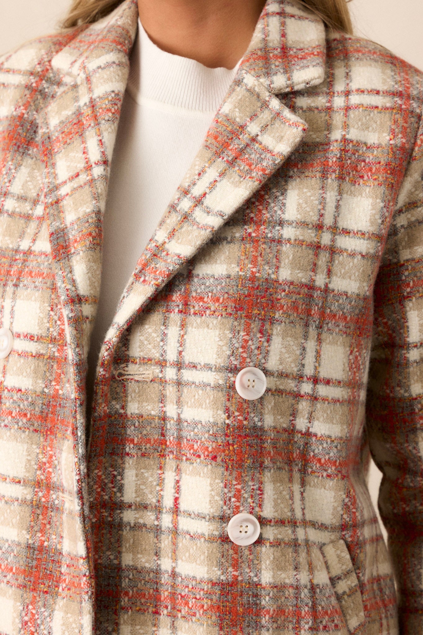 Close up view of this coat that features a collared neckline, double breasted buttons on the front, and a plaid pattern in shades of orange and beige.