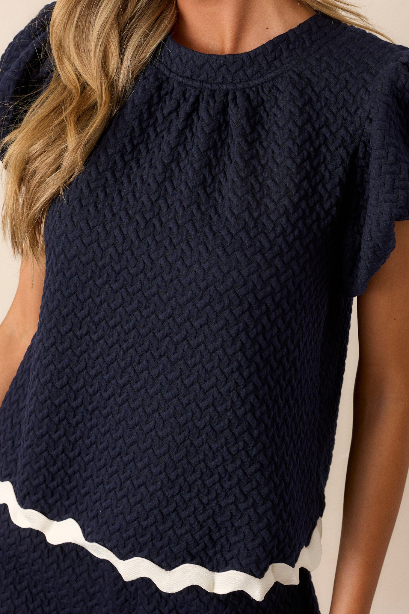 Close-up of the navy top showing the high rounded neckline, quilted material, and contrasting scalloped hemline.