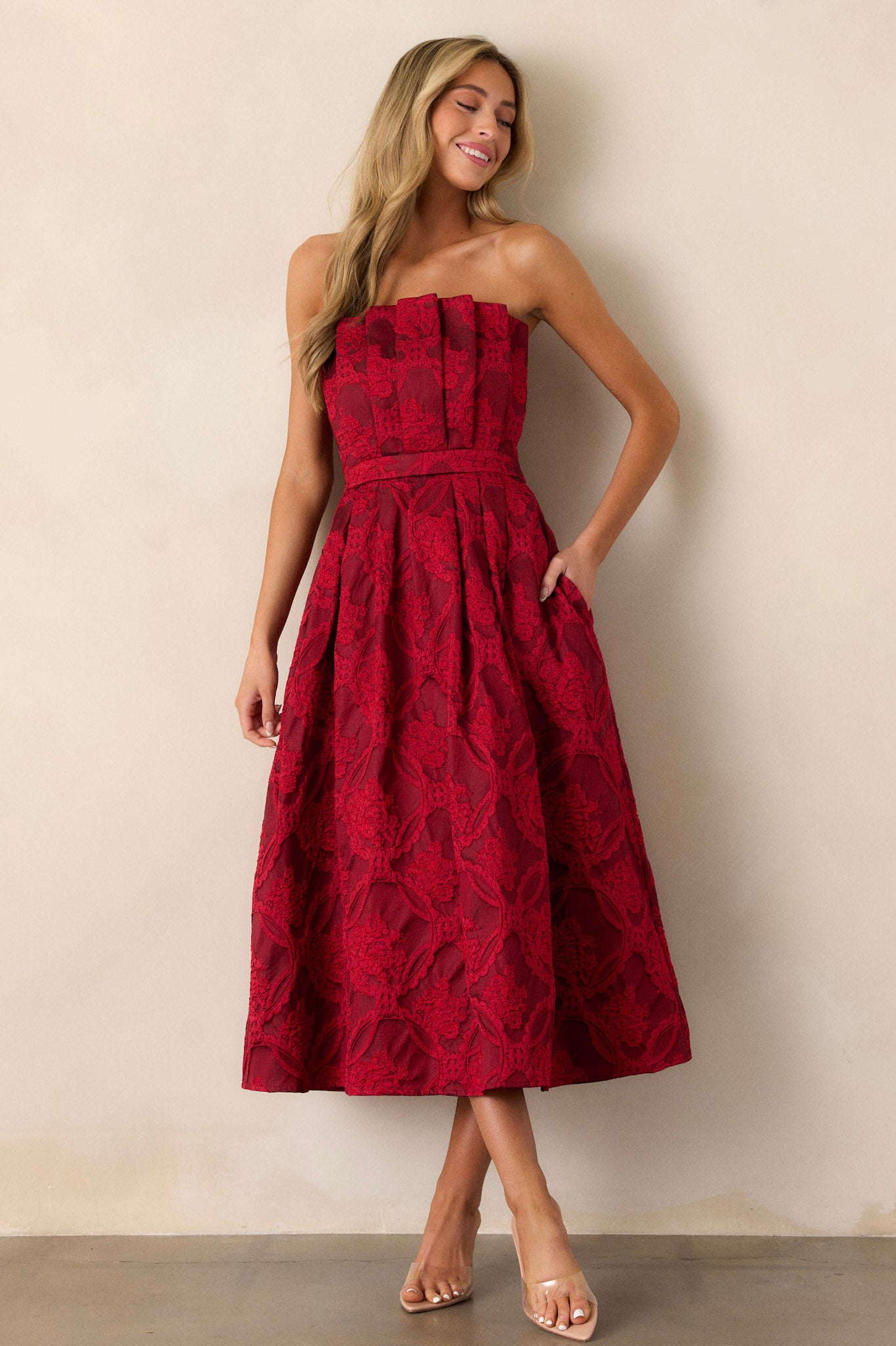 Full view of a red midi dress featuring a pleated bodice, a slightly pleated skirt, a jacquard design, functional hip pockets, a smocked back, a discrete back zipper and a strapless design.