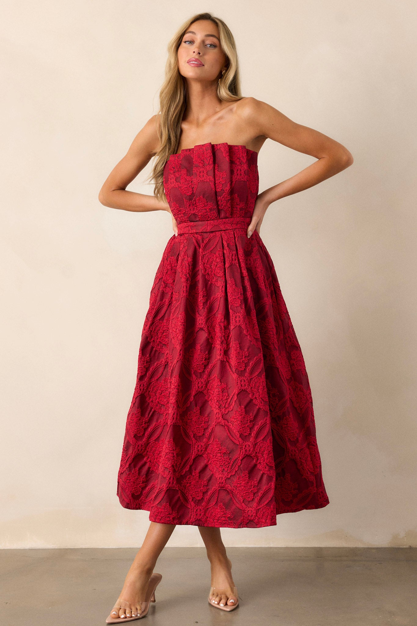 Side view of a red midi dress featuring a pleated bodice, a slightly pleated skirt, a jacquard design, functional hip pockets, a smocked back, a discrete back zipper and a strapless design.