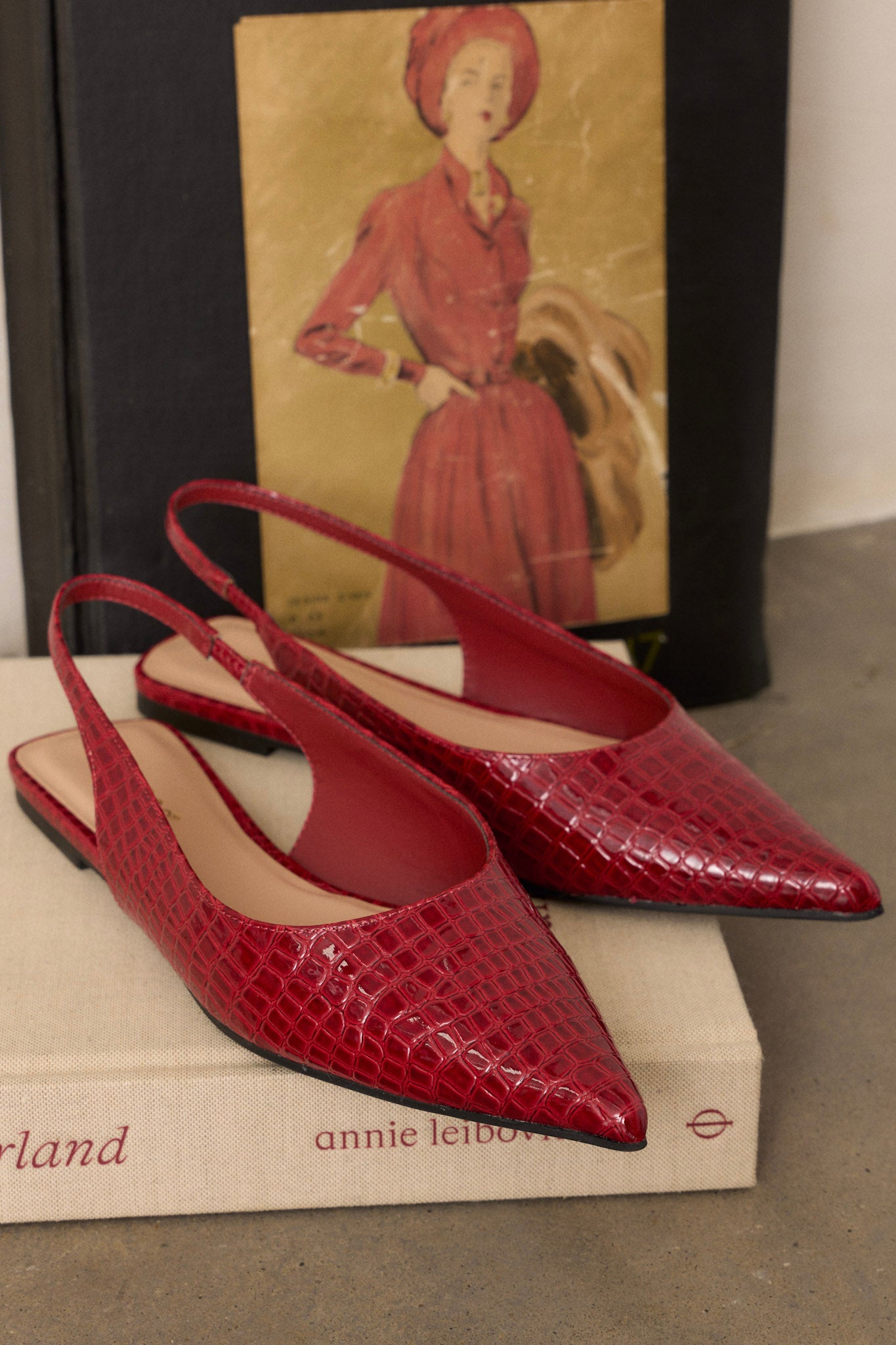 Stylized view of these red flats featuring a pointed toe, a faux crocodile leather, a slingback-style strap, and a sleek red color.