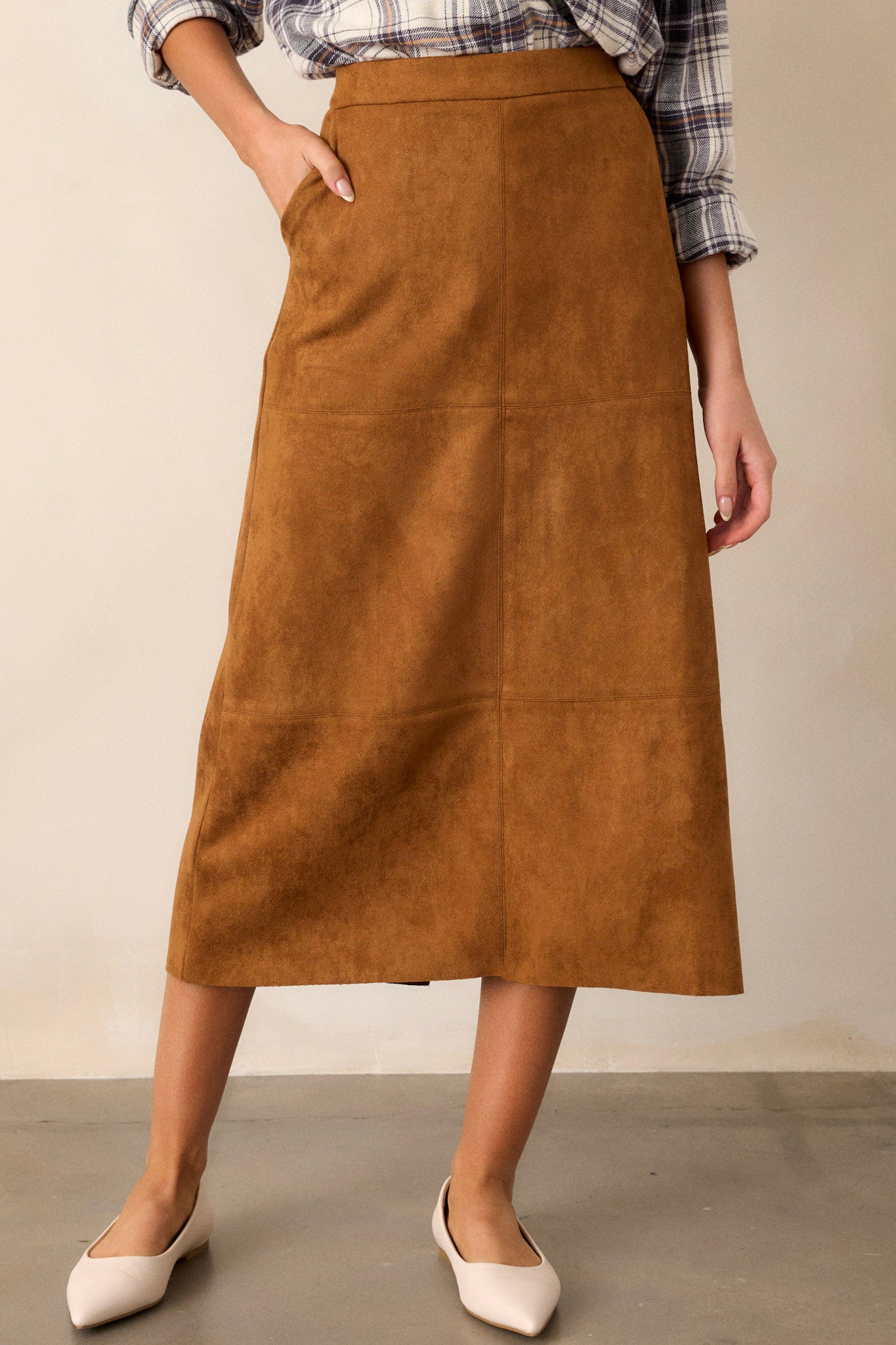 Front view of a midi skirt featuring a caramel brown faux suede material, an A-line silhouette, functional pockets, and a small slit up the back of the skirt.