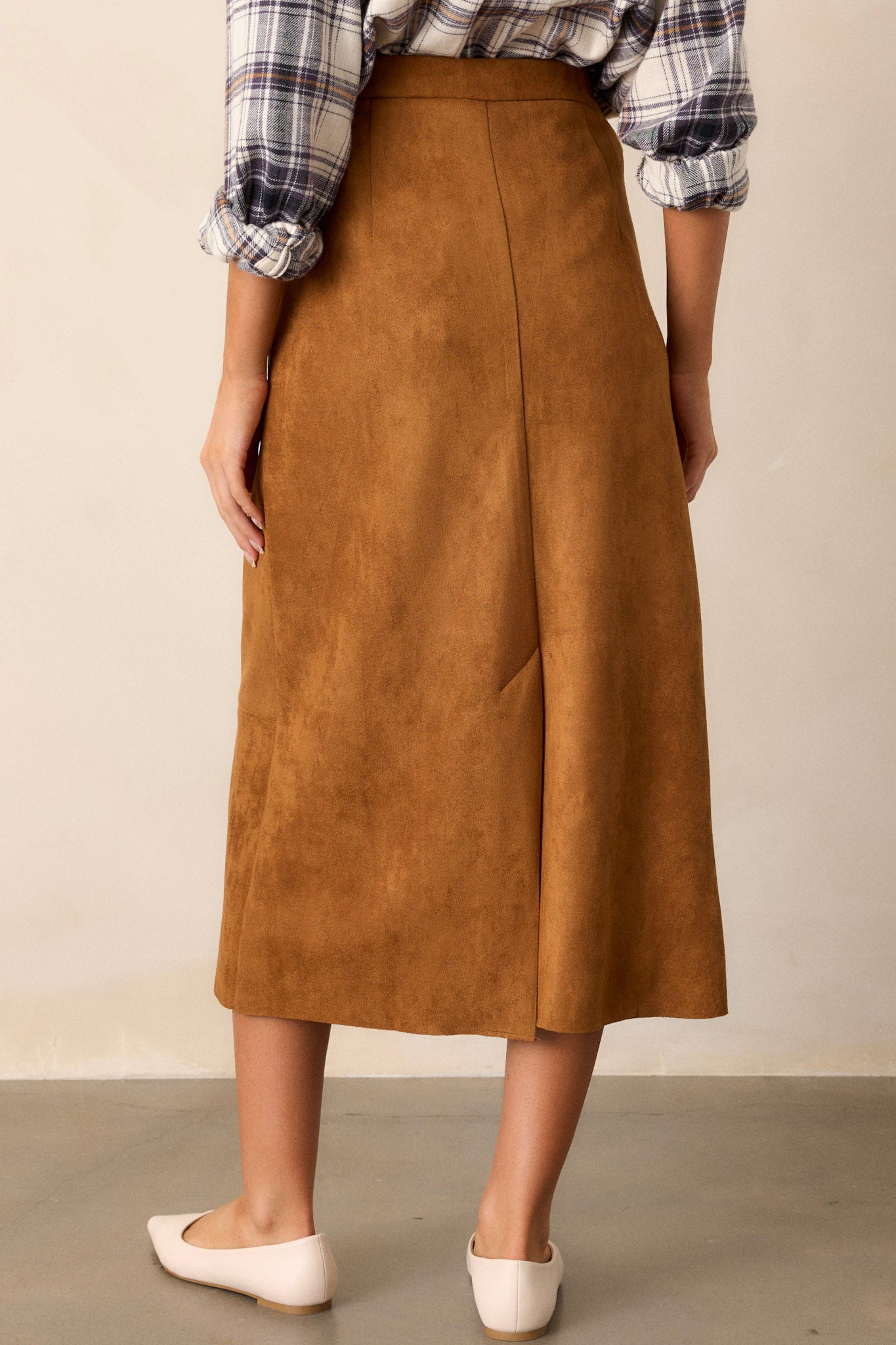 Back view of a midi skirt featuring a caramel brown faux suede material, an A-line silhouette, functional pockets, and a small slit up the back of the skirt.