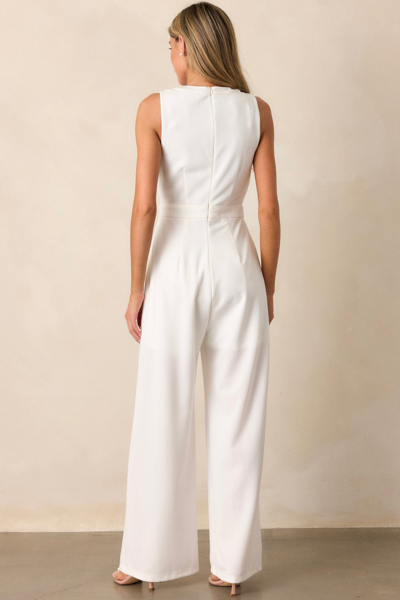 Back view of this white jumpsuit that features a round neckline, a discrete back zipper, three ornate bows embellished with faux ivory pearls and rhinestones, a chest cutout, a thick waistband, subtle pleats, functional hip pockets, and a wide leg design.