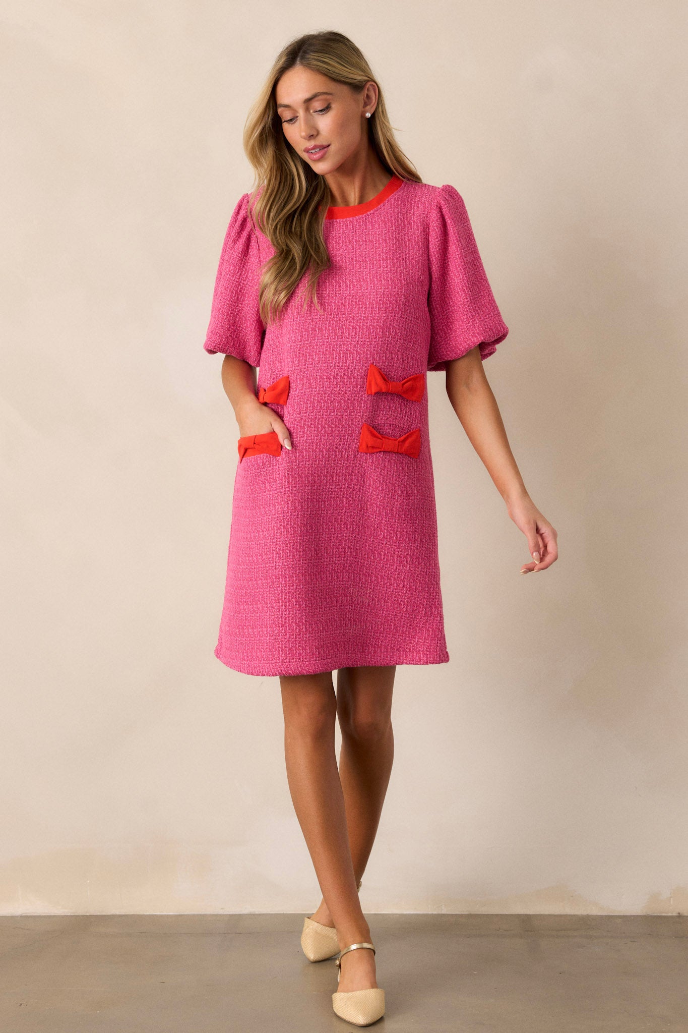 This pink tweed dress features a high rounded neckline, a relaxed fit, red bow detailing on the hips, functional hip pockets, a discrete back zipper and elastic cuffed short sleeves.