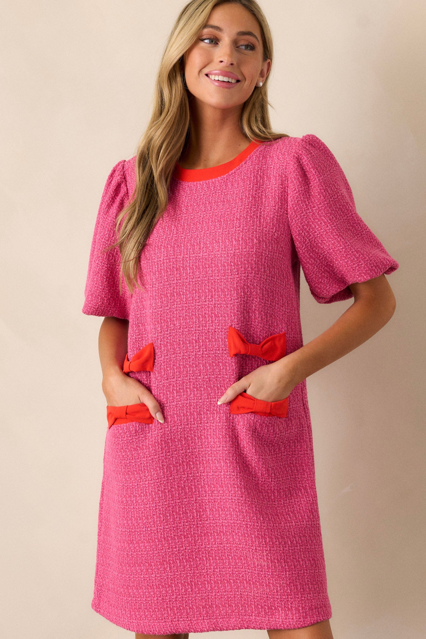 Full length view of a pink tweed dress featuring a high rounded neckline, red bow detailing on the hips, functional hip pockets, a discrete back zipper, and elastic cuffed short sleeves.