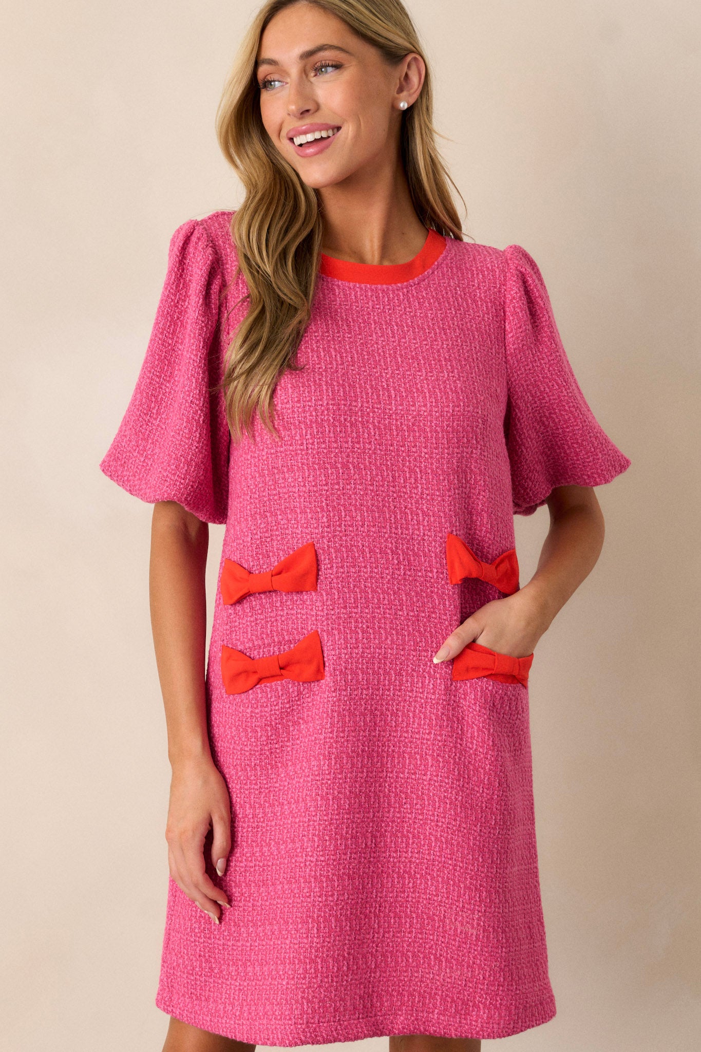 Front angled view of a pink tweed dress featuring a high rounded neckline, red bow detailing on the hips, functional hip pockets, a discrete back zipper, and elastic cuffed short sleeves.