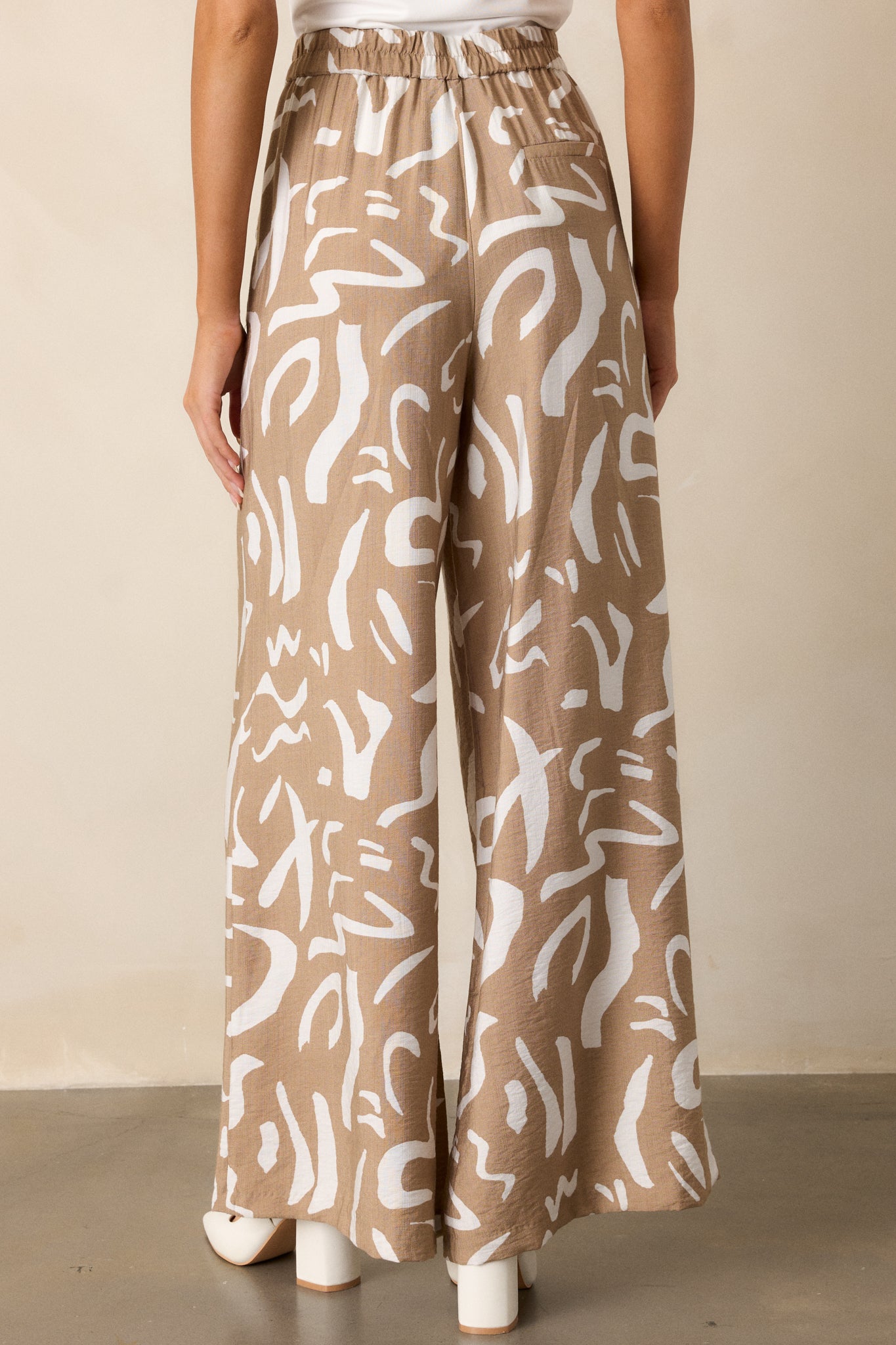 Rear view of high-waisted camel pants with a wide leg fit, featuring the button and zipper closure and abstract pattern across the fabric.