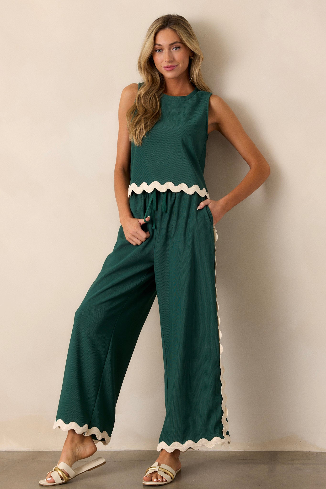 Full body view of these green pants featuring a high waisted design, an elastic waistband, a self-tie drawstring, functional hip pockets, a ribbed texture, and ricrac hemlines.