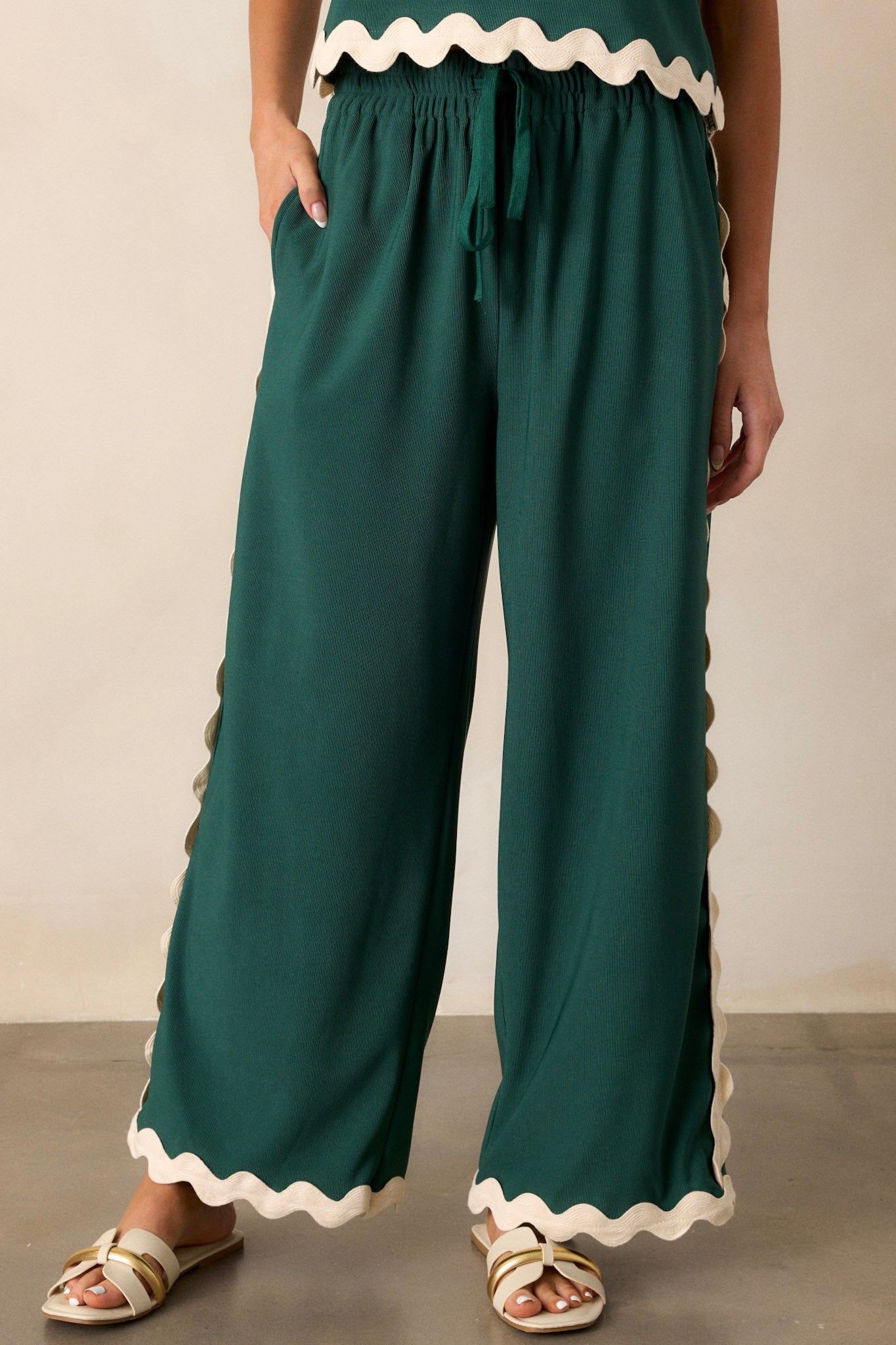 Front view of green pants featuring a high waisted design, an elastic waistband, a self-tie drawstring, functional hip pockets, a ribbed texture, and ricrac hemlines.