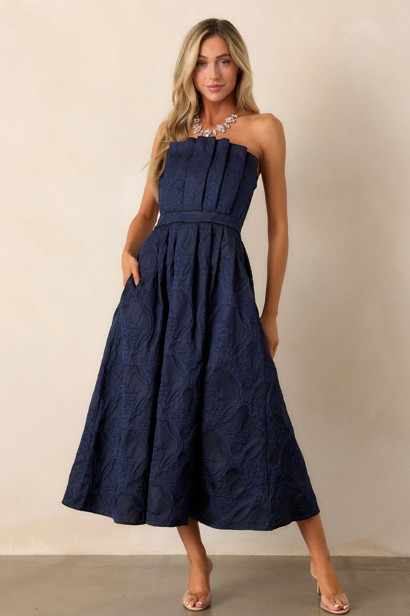 Front view of a navy midi dress featuring a pleated bodice, a slightly pleated skirt, a jacquard design, functional hip pockets, a smocked back, a discrete back zipper and a strapless design.