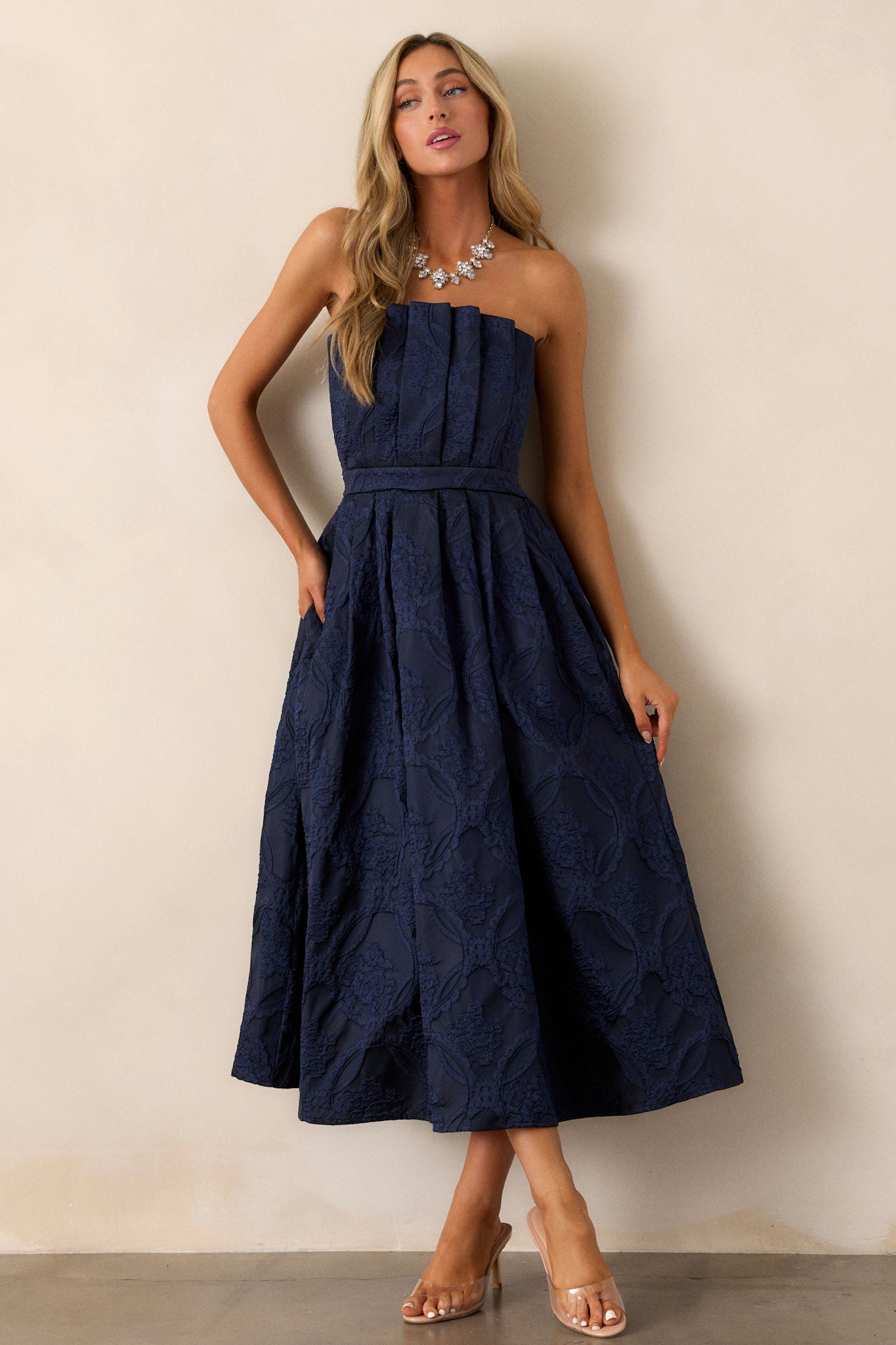 Full view of a navy midi dress featuring a pleated bodice, a slightly pleated skirt, a jacquard design, functional hip pockets, a smocked back, a discrete back zipper and a strapless design.