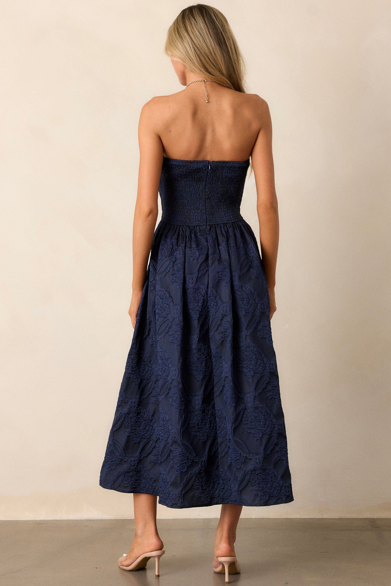 Back view of a navy midi dress featuring a pleated bodice, a slightly pleated skirt, a jacquard design, functional hip pockets, a smocked back, a discrete back zipper and a strapless design.