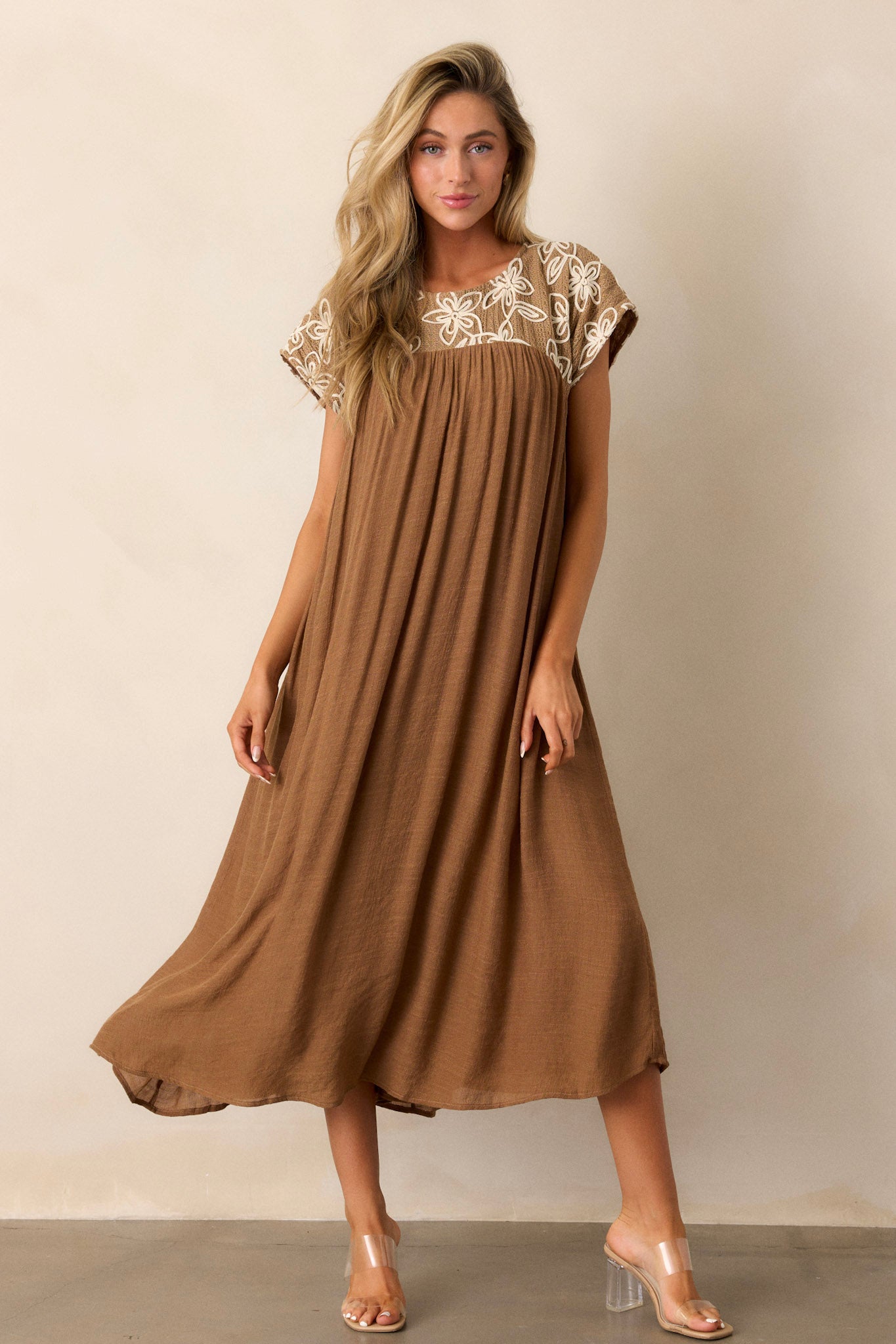 Tan midi dress highlighting the high rounded neckline, embroidered floral pattern, and short sleeves with a flowing silhouette.