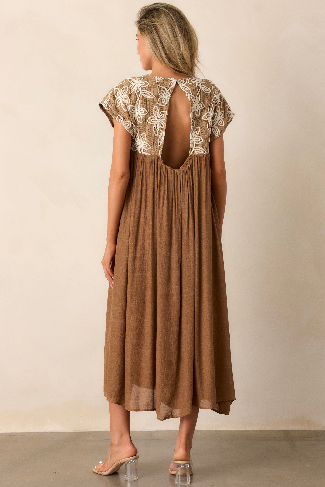 Back view of the tan midi dress highlighting the high rounded neckline, embroidered floral pattern, and flowing silhouette.