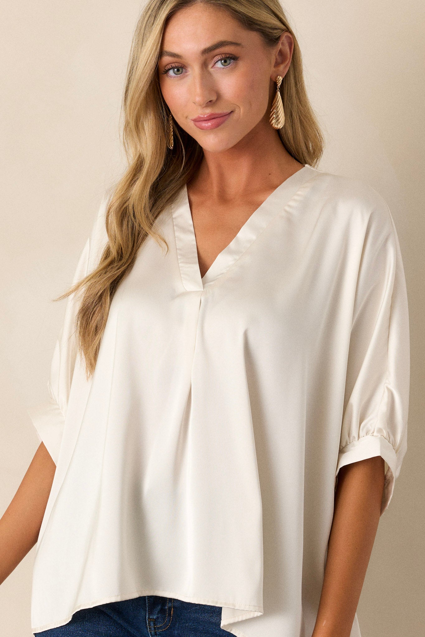 Front angled view of a white top featuring a v-neckline, short dolman sleeves with pleated detailing, a wide, flowy fit, and a satin-like material