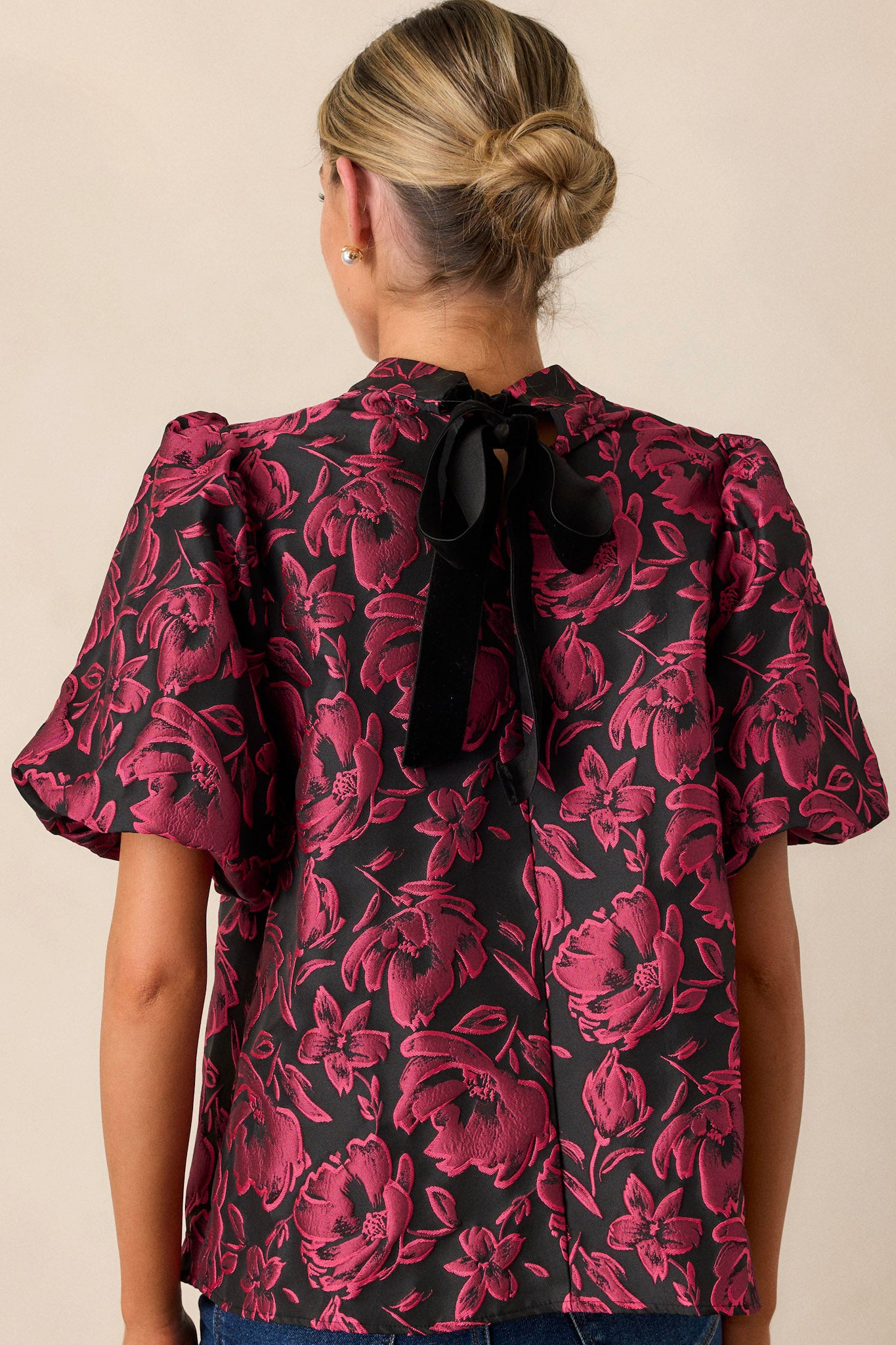 A view of the back of the dark fuchsia top, highlighting the mock neckline and smooth continuation of the floral jacquard fabric without any detailing from the bow.