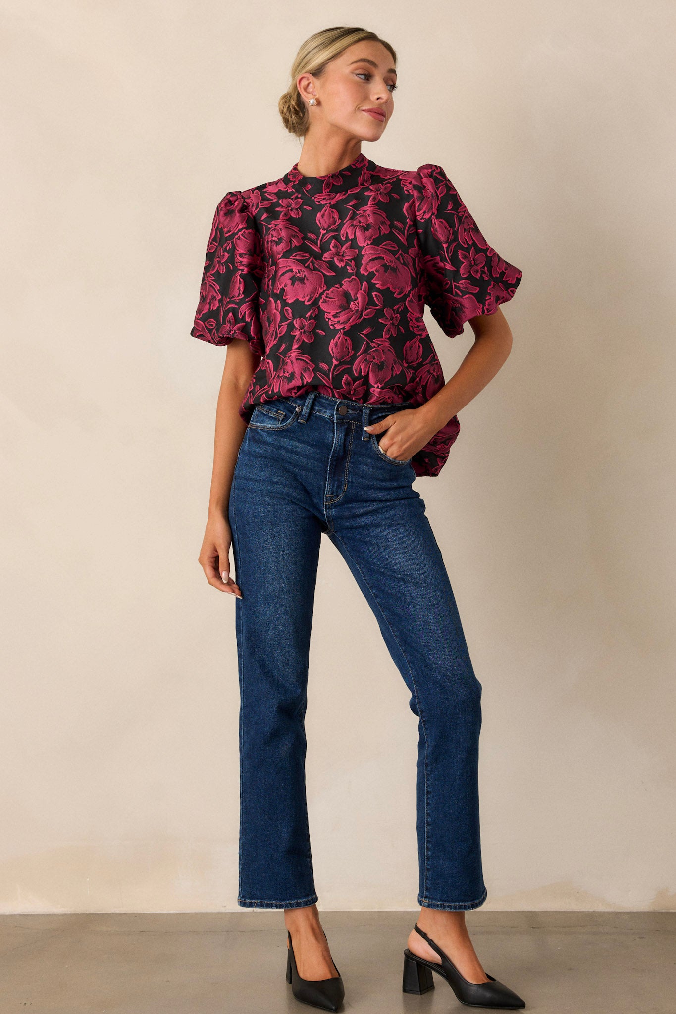 A dark fuchsia top with a mock neckline, short puff sleeves, and floral jacquard fabric, featuring a faux velvet self-tie bow at the neck, shown in full-length view.