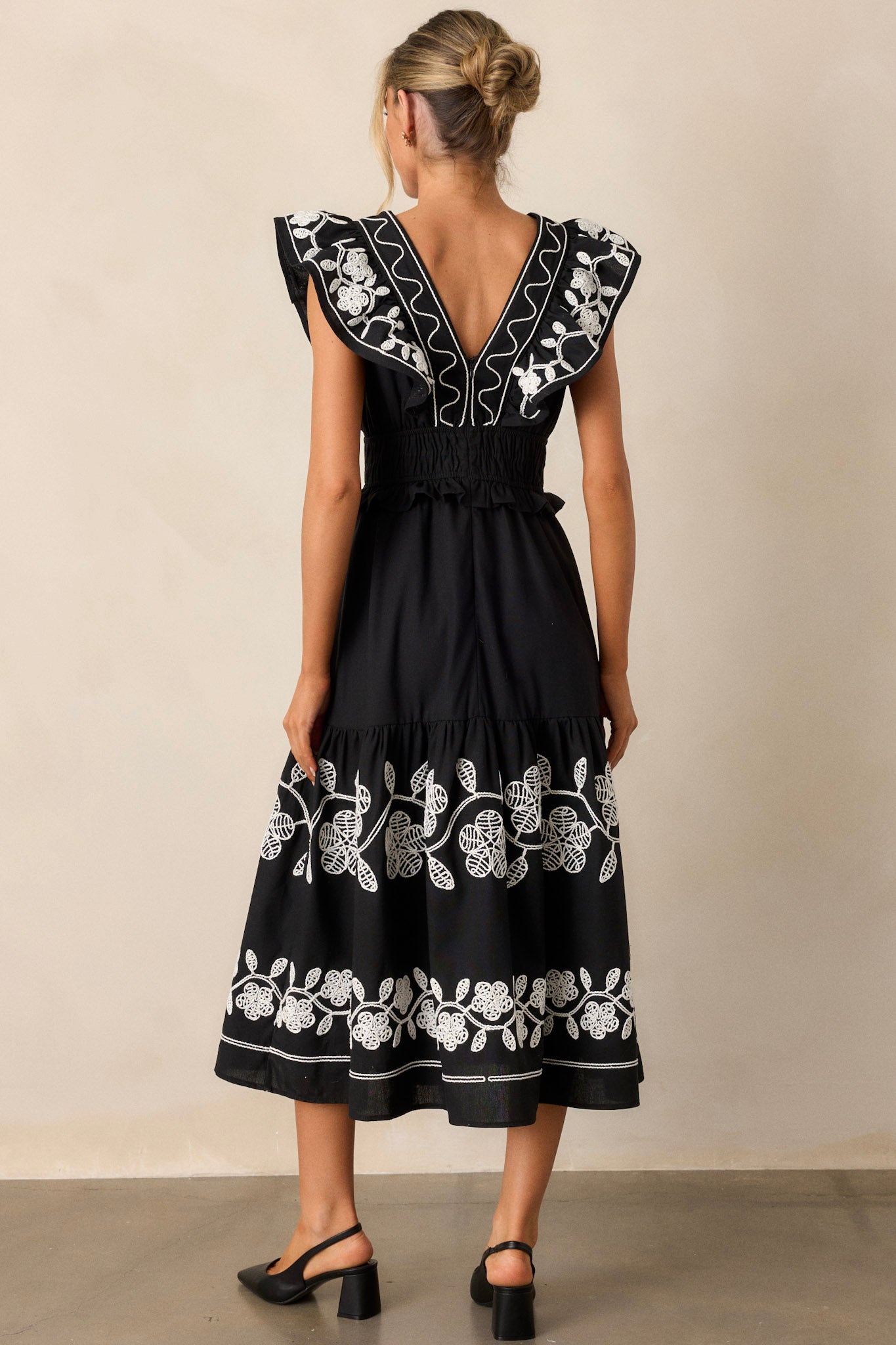 Back view of the black dress highlighting the smocked waist, the discrete back zipper, and the flow of the short flutter sleeves, with embroidered detailing visible across the fabric.