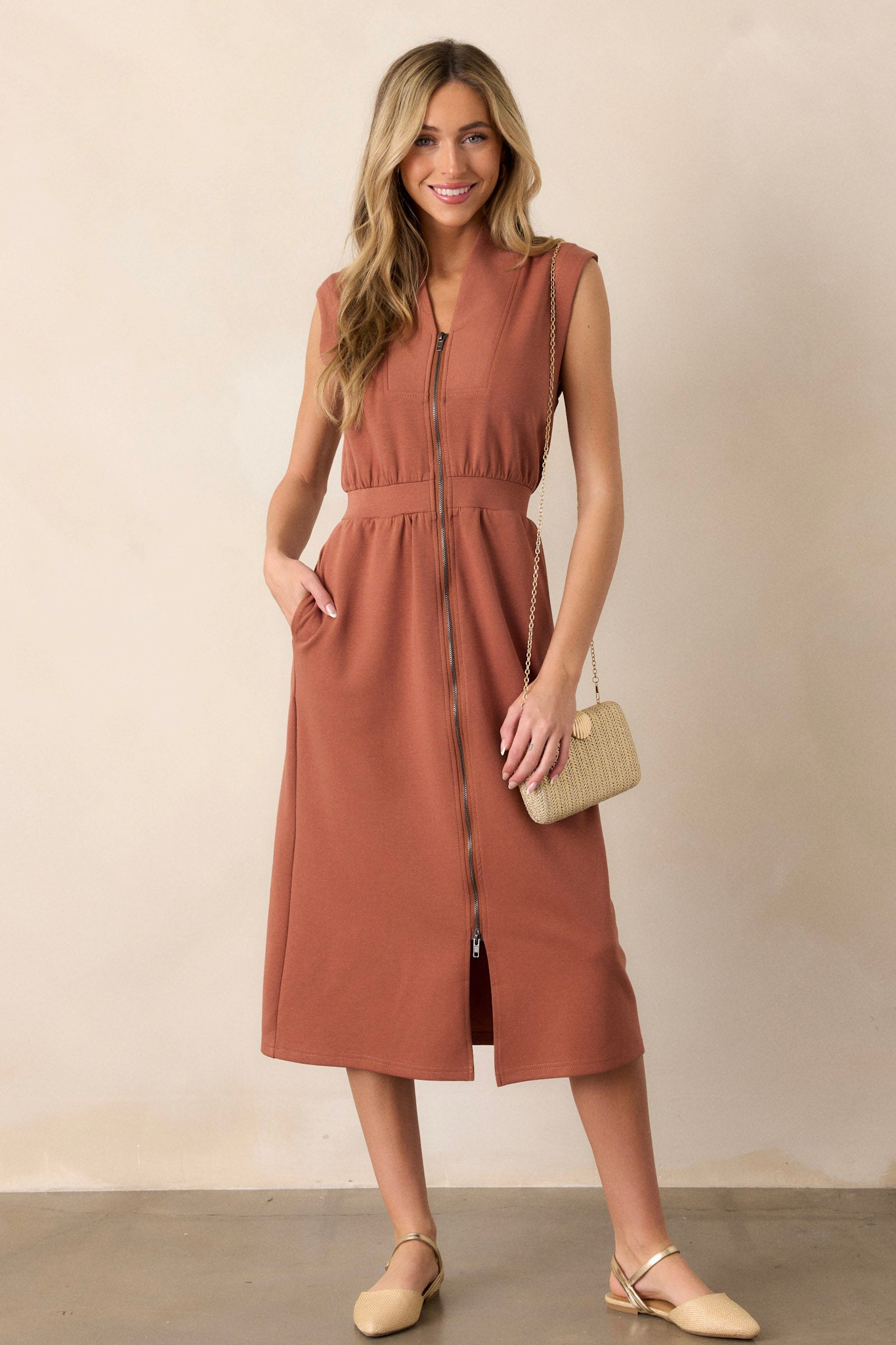 Front view of this terracotta dress that features a v-neckline, a slight collar, a functional full zip front, functional hip pockets, and a sleeveless design.