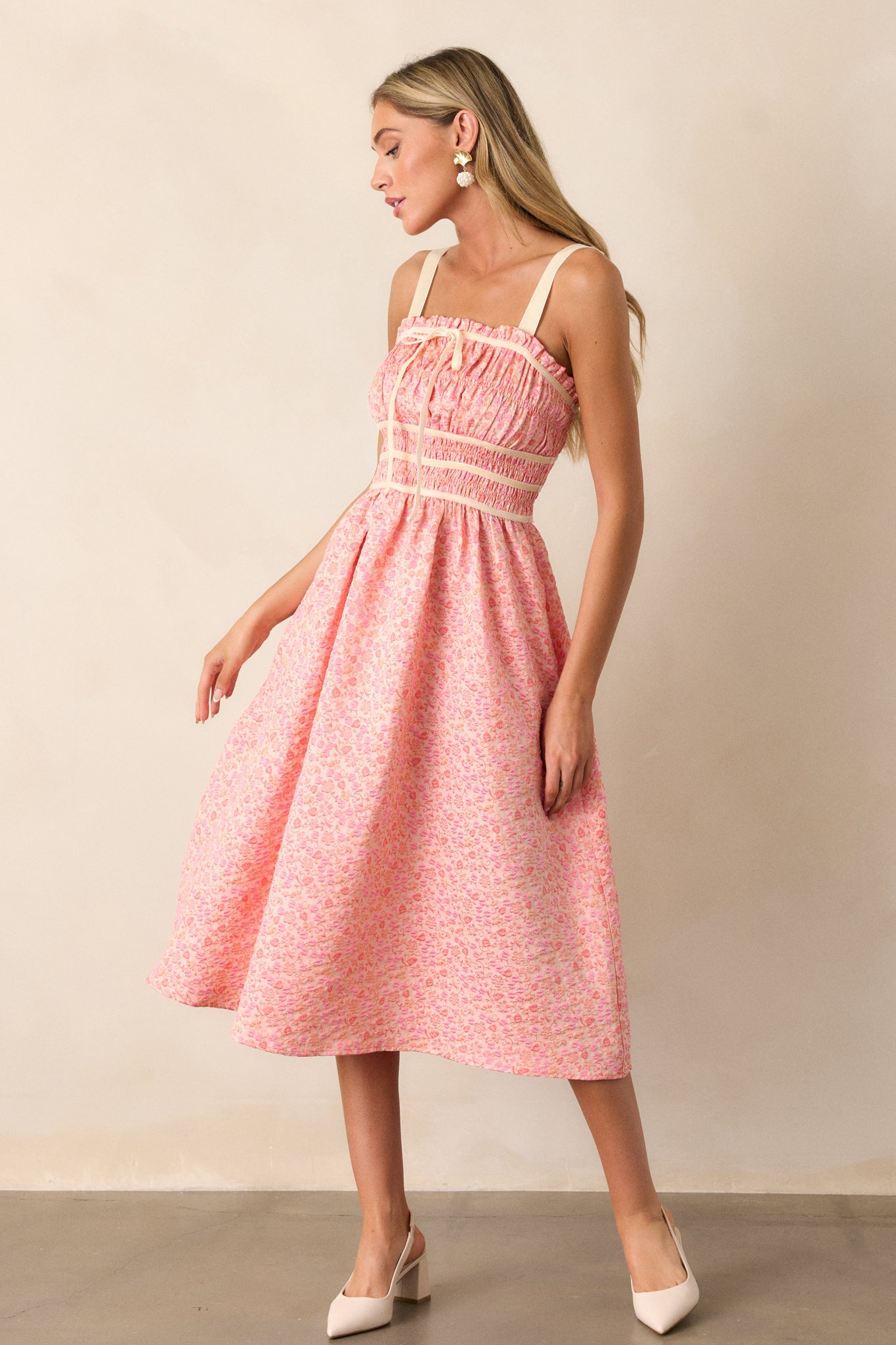 This pink coral midi dress features a square neckline, thick adjustable straps, fully smocked bust and waist, a self-tie bust feature, velvet accents, functional hip pockets, and embossment throughout.