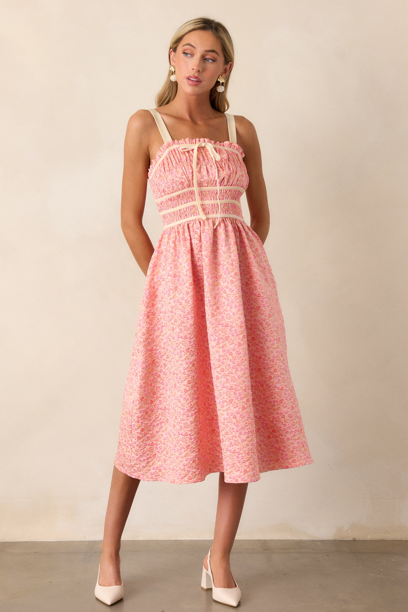 Front view of this pink coral midi dress that features a square neckline, thick adjustable straps, fully smocked bust and waist, a self-tie bust feature, velvet accents, functional hip pockets, and embossment throughout.